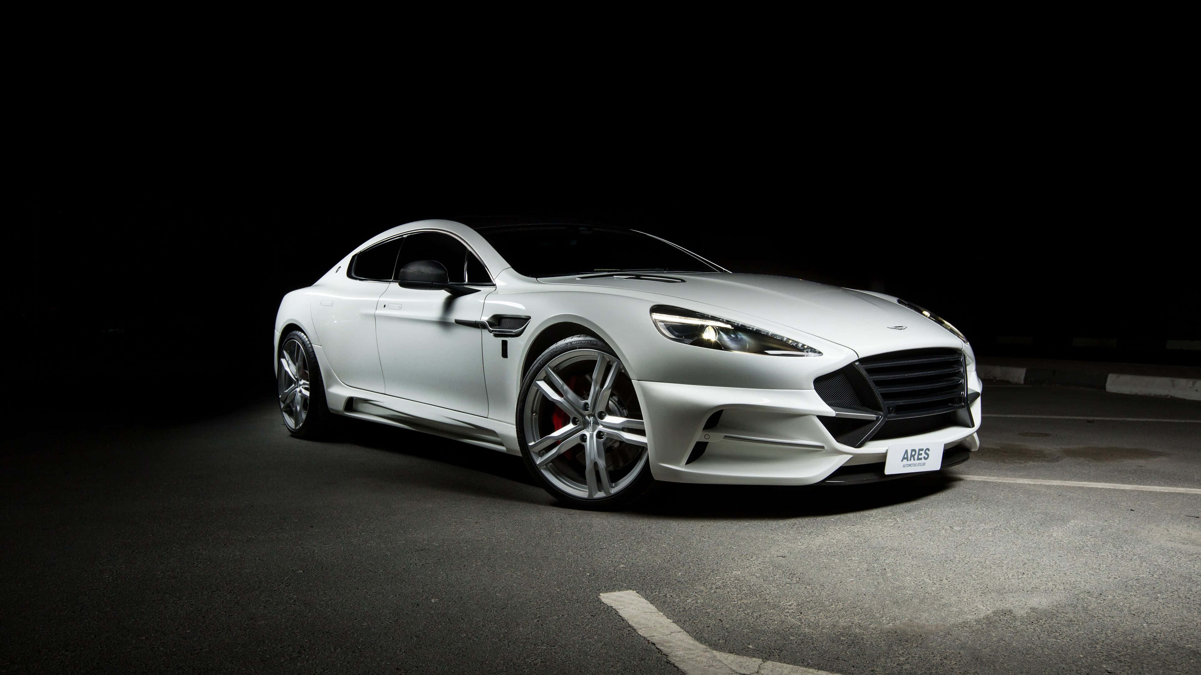 Aston Martin Rapide S 桌布 壁紙 — 一架流線型白色Aston Martin Ares跑車在漆黑夜晚的停車場中，於戲劇性的聚光燈下閃閃發光。