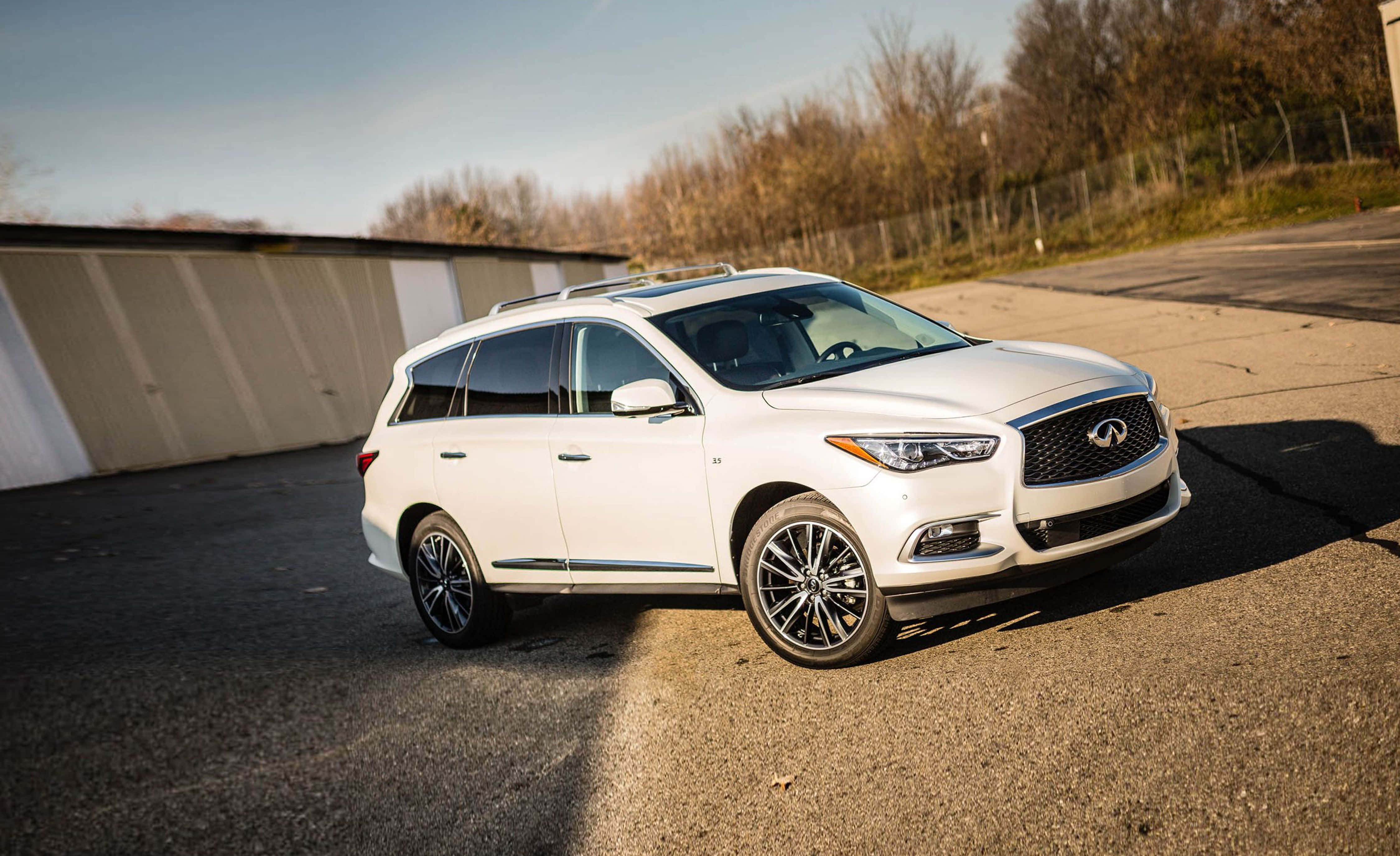 Infiniti Qx60 桌布 壁紙 — 一架流線型白色Infiniti QX60 SUV停泊在瀝青路上，背景有光禿樹木和倉庫，天空晴朗。