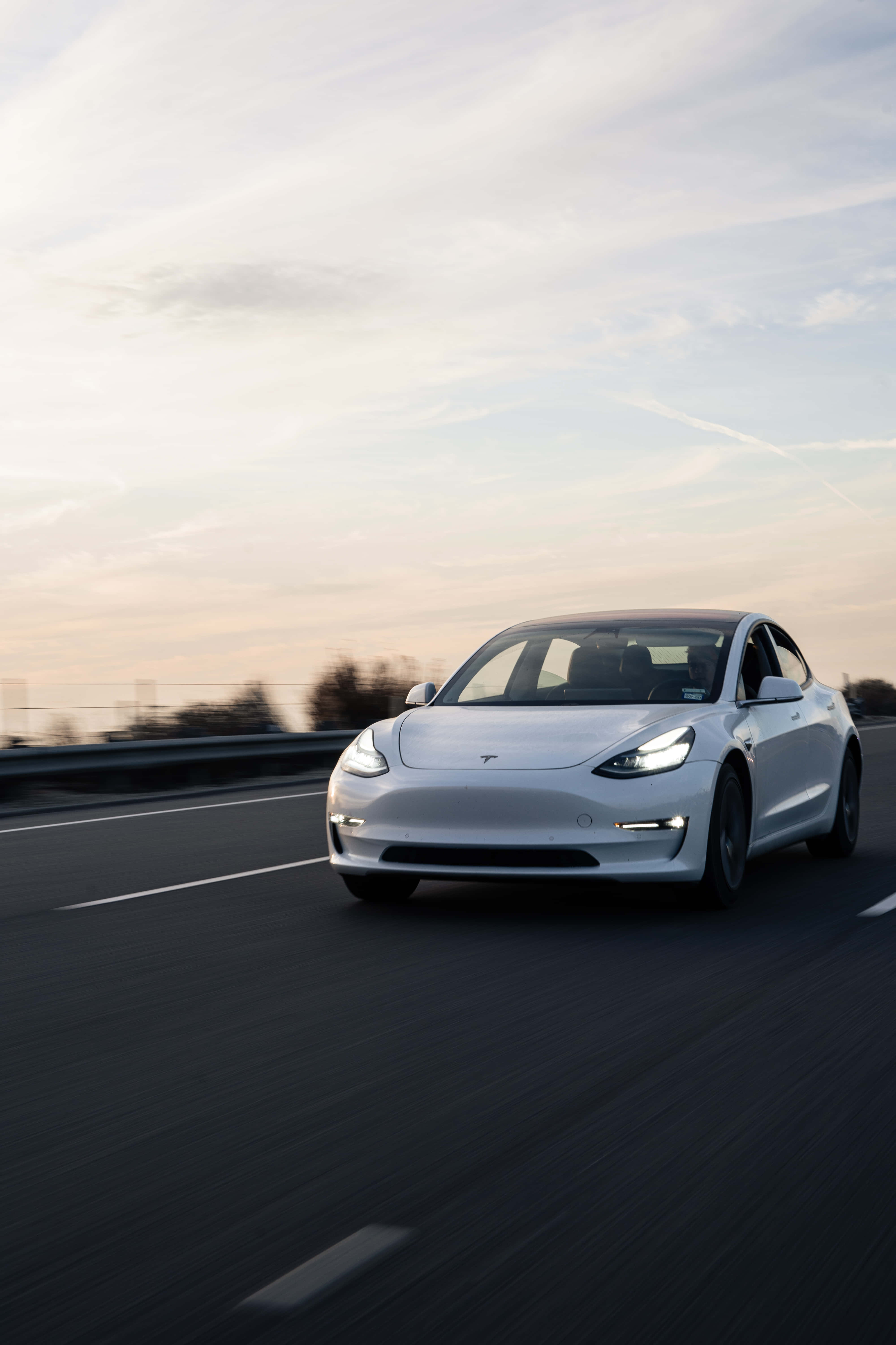 tesla 桌布 壁紙 — 一架流線型白色Tesla Model 3在柔和粉彩夕陽天空下沿高速公路飛馳，帶有模糊的動態線條。