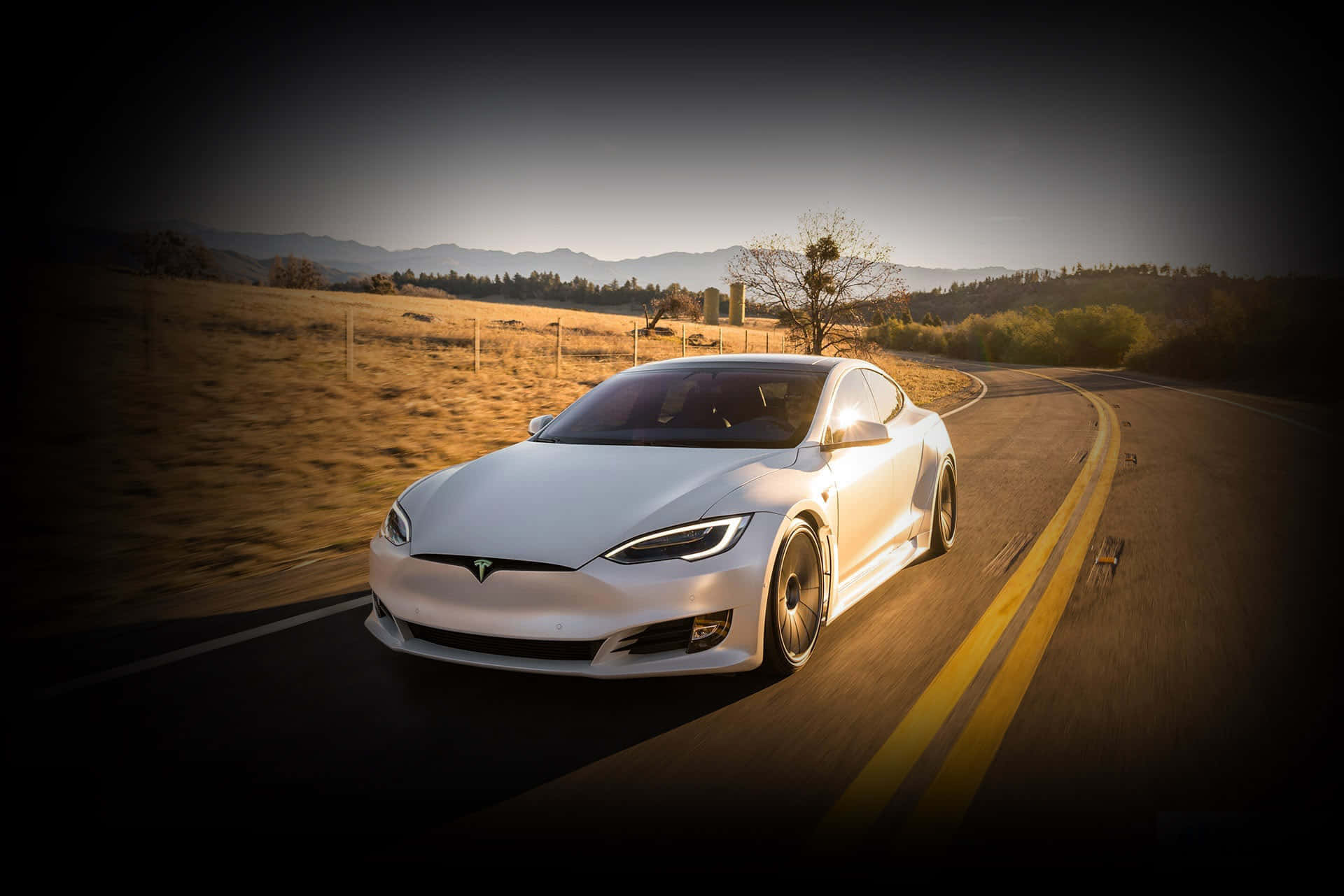tesla 桌布 壁紙 — 一架流線型白色Tesla Model S在黃金時段沿著蜿蜒的鄉村道路行駛，遠處有山巒。