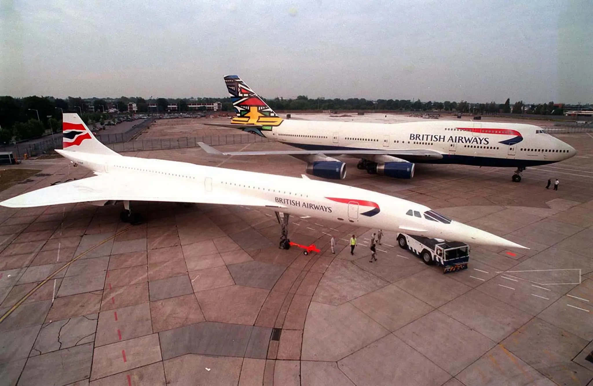 Jumbo Jets 桌布 壁紙 — 一架流線型白色和諧客機與一架英國航空波音747並排停在停機坪上，附近有地勤人員。