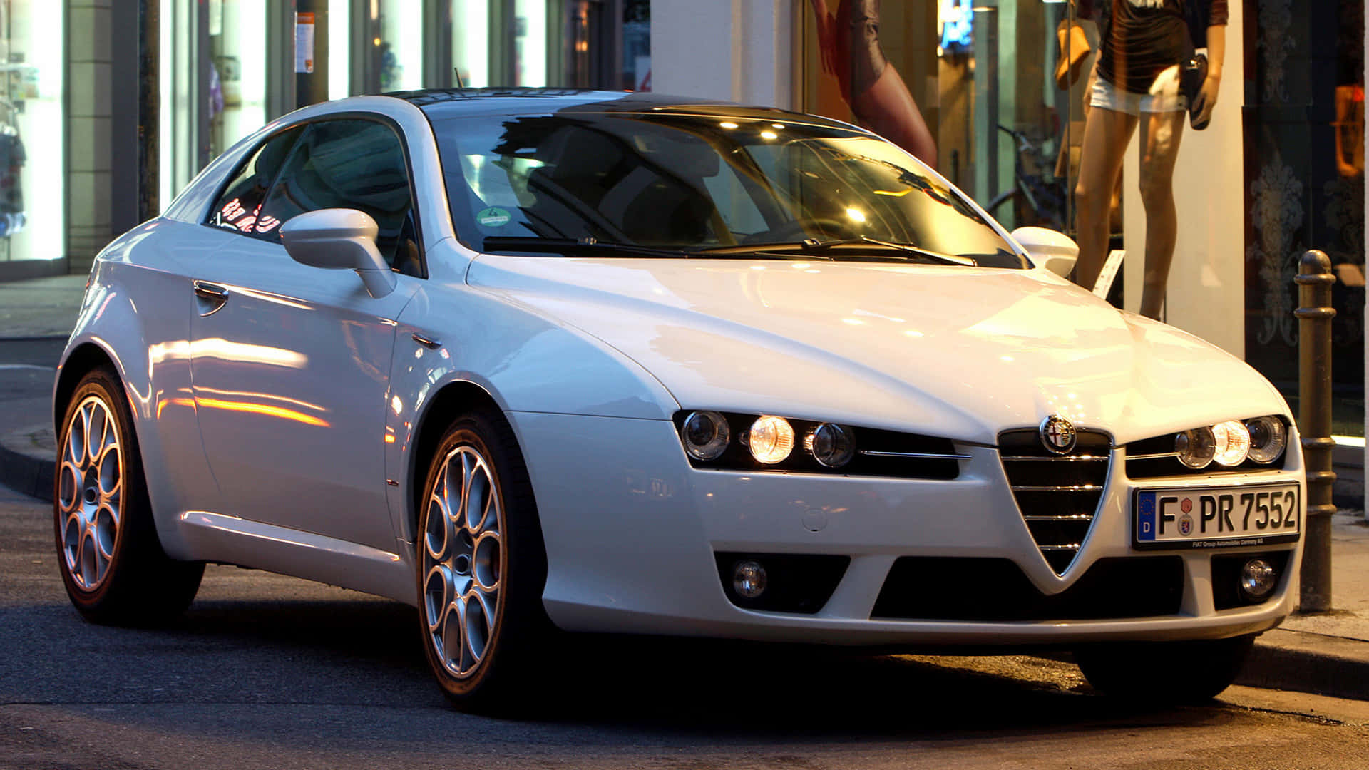 Alfa Romeo Brera 桌布 壁紙 — 一架流線型白色愛快羅密歐跑車在金色街燈下閃閃發光，停泊在商店櫥窗附近的城市行人路上。