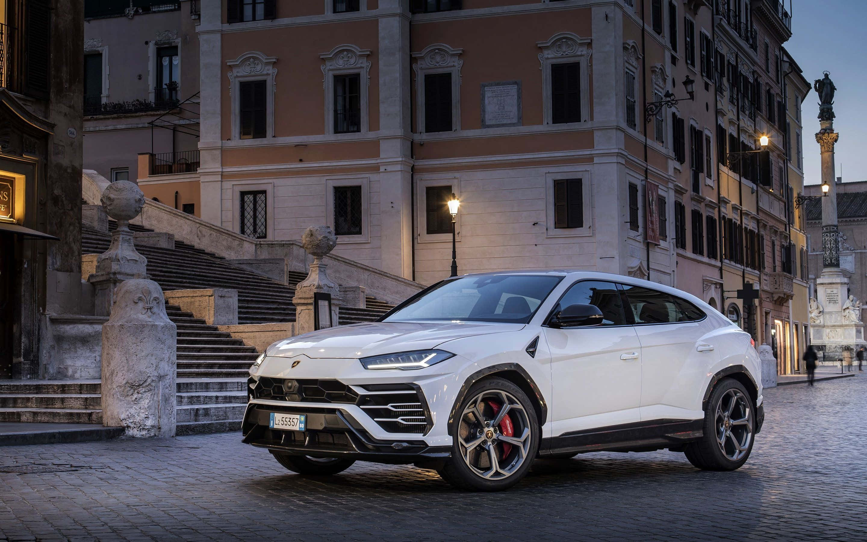 Lamborghini Urus 桌布 壁紙 — 一架流線型白色林寶堅尼Urus SUV停泊在黃昏的鵝卵石街道上，背景是歷史悠久的歐洲建築和溫暖的路燈。