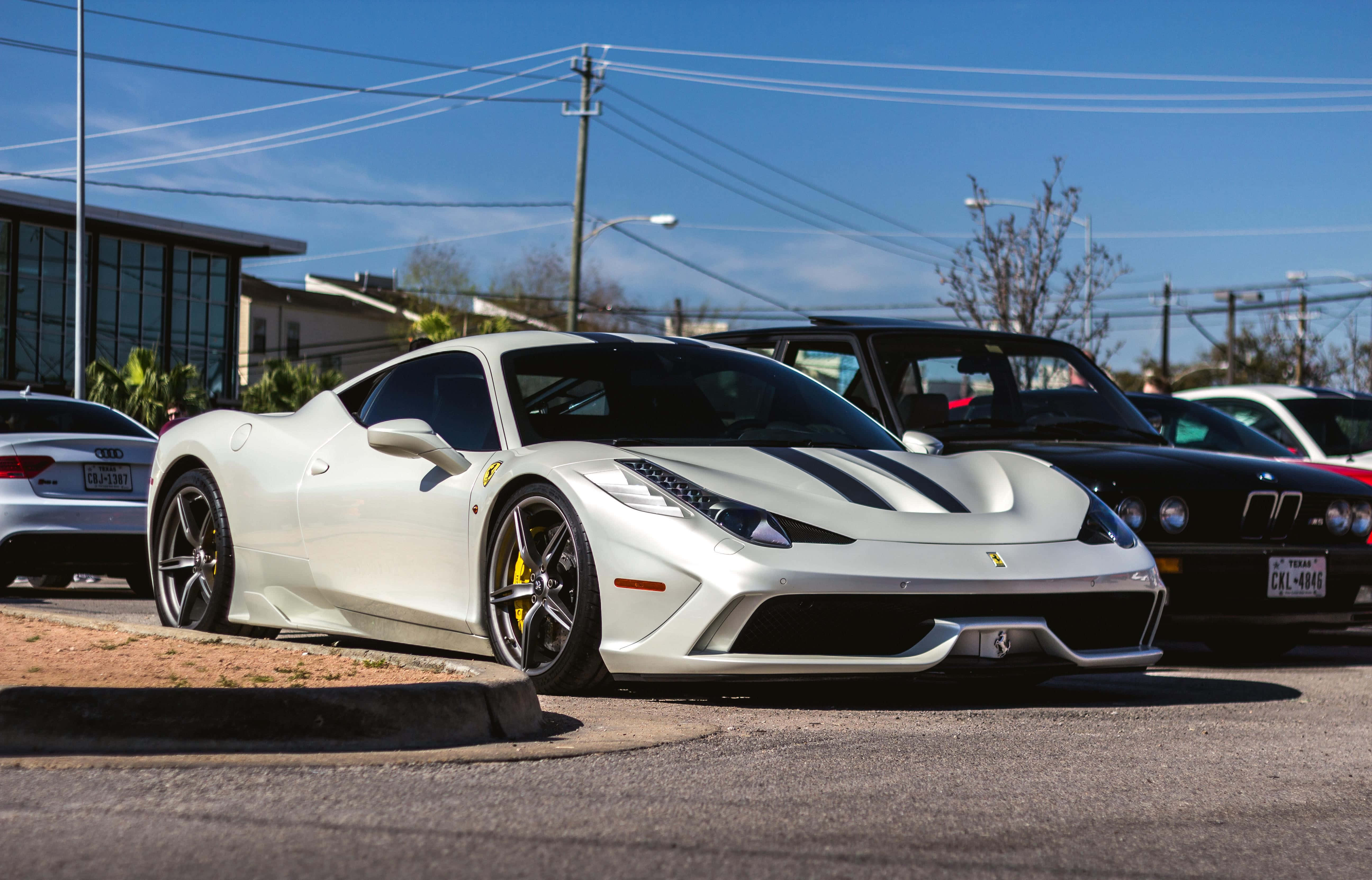 Ferrari 458 Speciale 桌布 壁紙 — 一架流線型白色法拉利跑車帶有黑色賽車條紋，停泊在陽光明媚的城市地段，旁邊有其他豪華車輛。