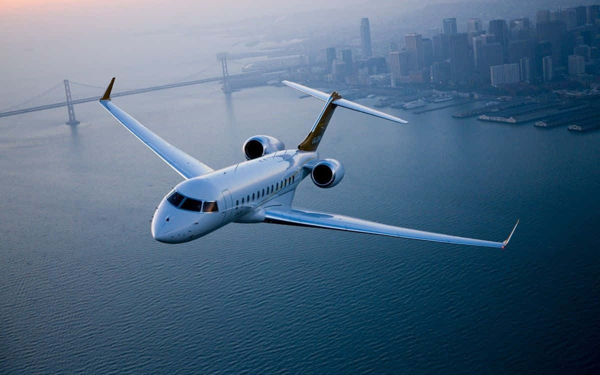 Jet 桌布 壁紙 — 一架流線型白色私人飛機在一片廣闊的藍色海灣上空翱翔，下方可見城市天際線和一座吊橋。