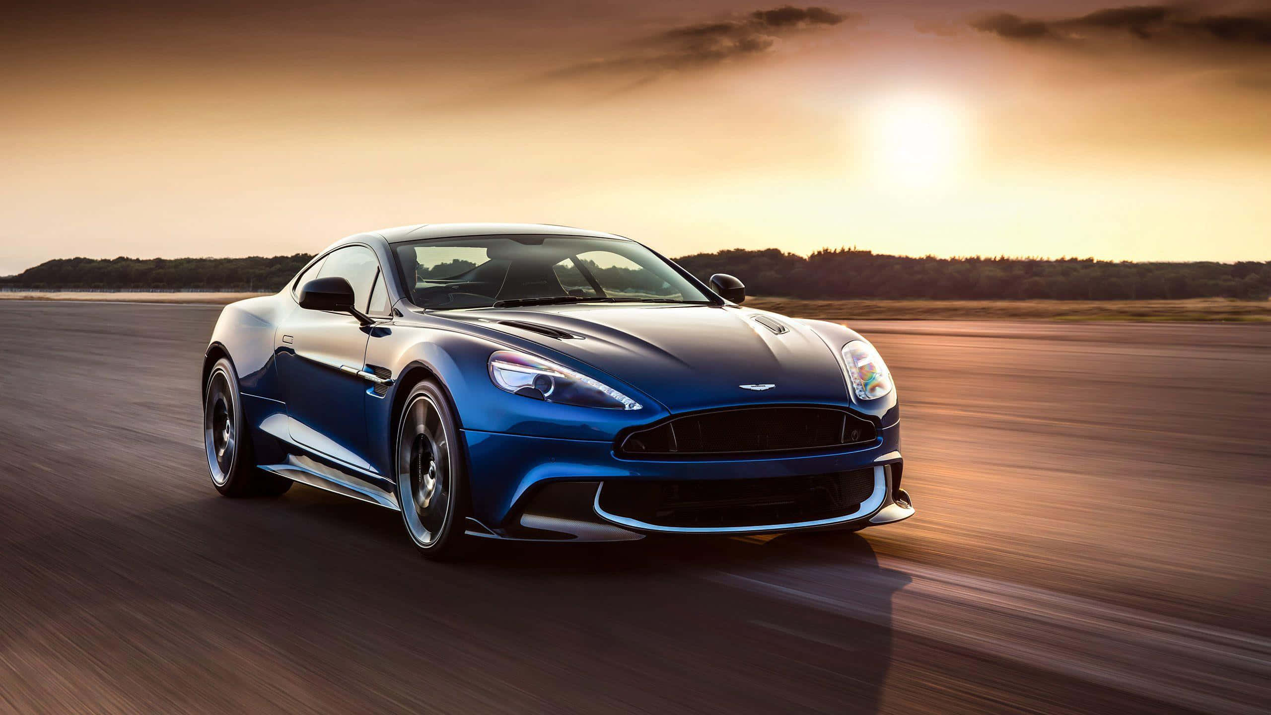 Aston Martin V12 Vantage 桌布 壁紙 — 一架流線型藍色Aston Martin在日落時分於跑道上疾馳，金色光線在模糊的瀝青上延伸。