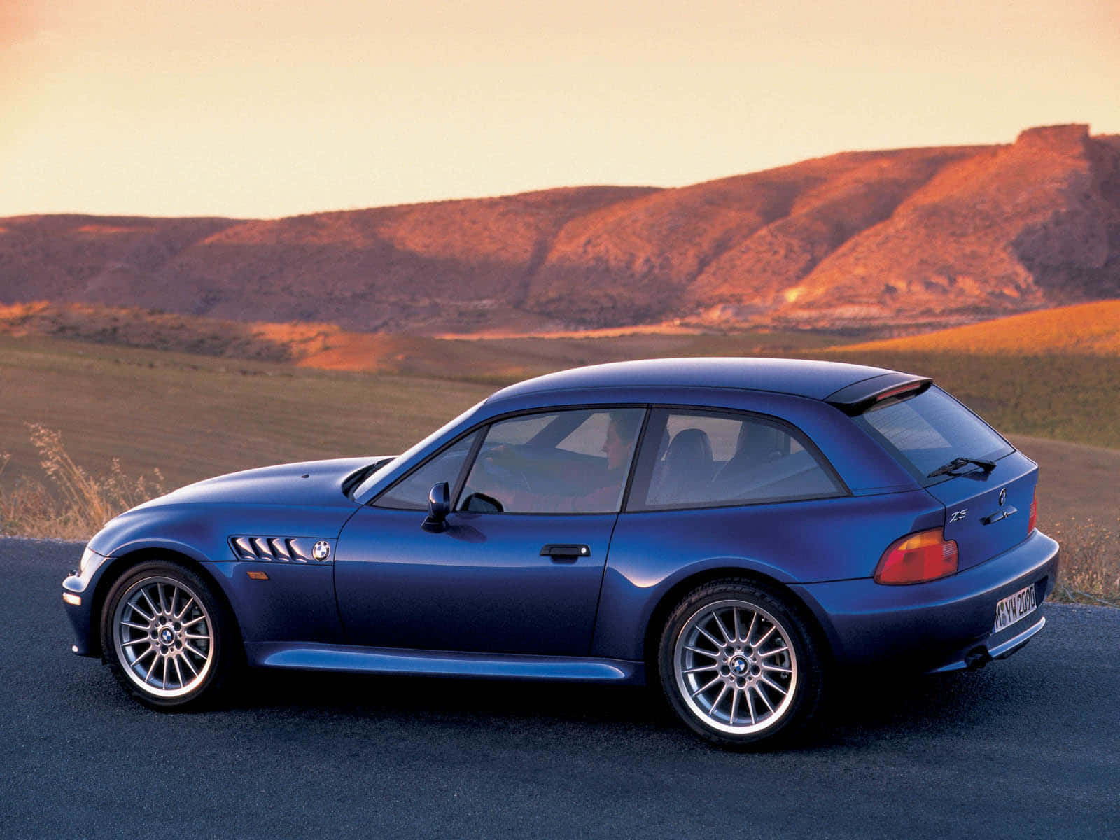 Bmw Z3 桌布 壁紙 — 一架流線型藍色BMW Z3開篷跑車停泊在沙漠公路上，背景是夕陽下的連綿山丘。
