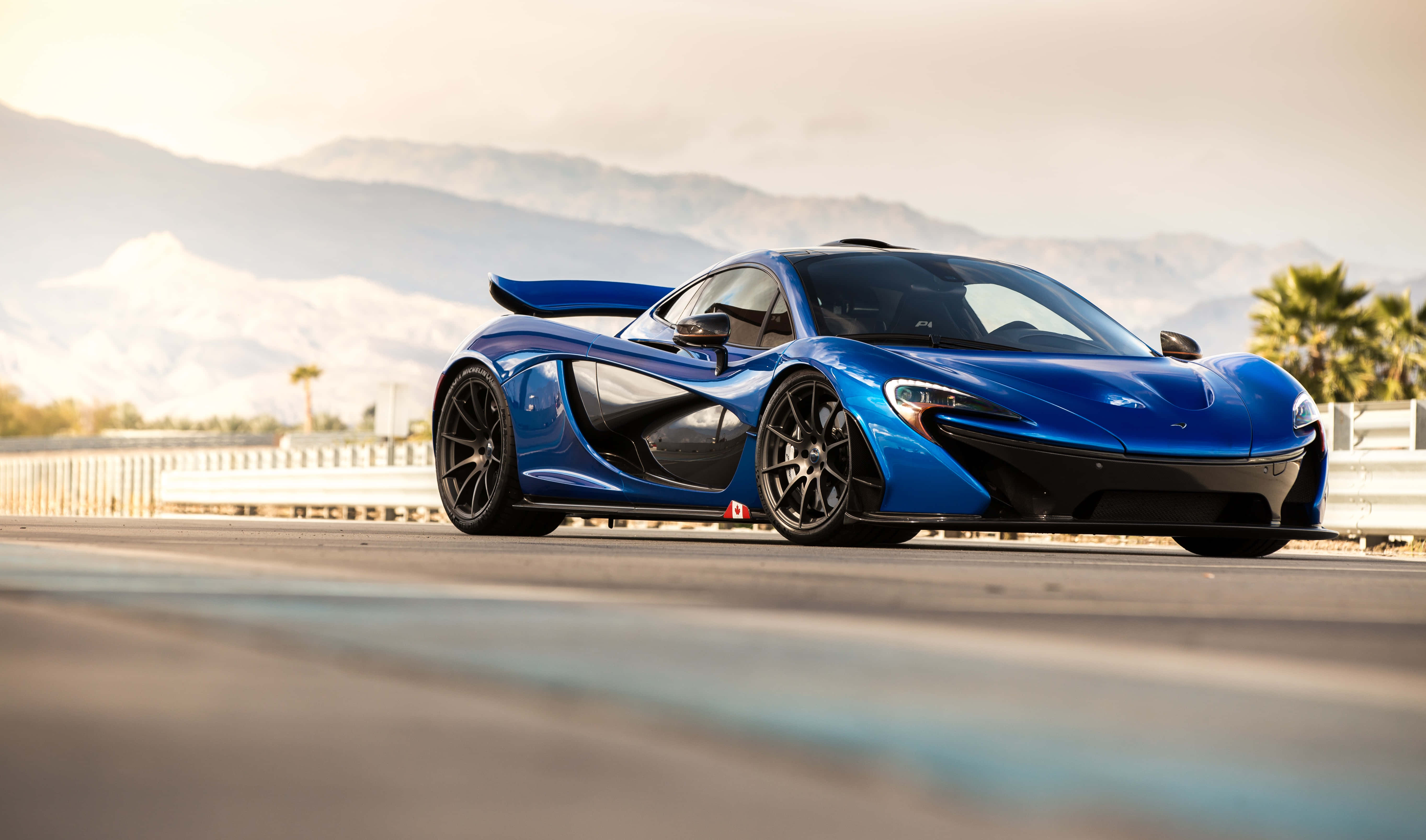 麥拿侖 桌布 壁紙 — 一架流線型藍色McLaren P1跑車在沙漠公路上滑行，遠方有山脈和棕櫚樹，天空朦朧。