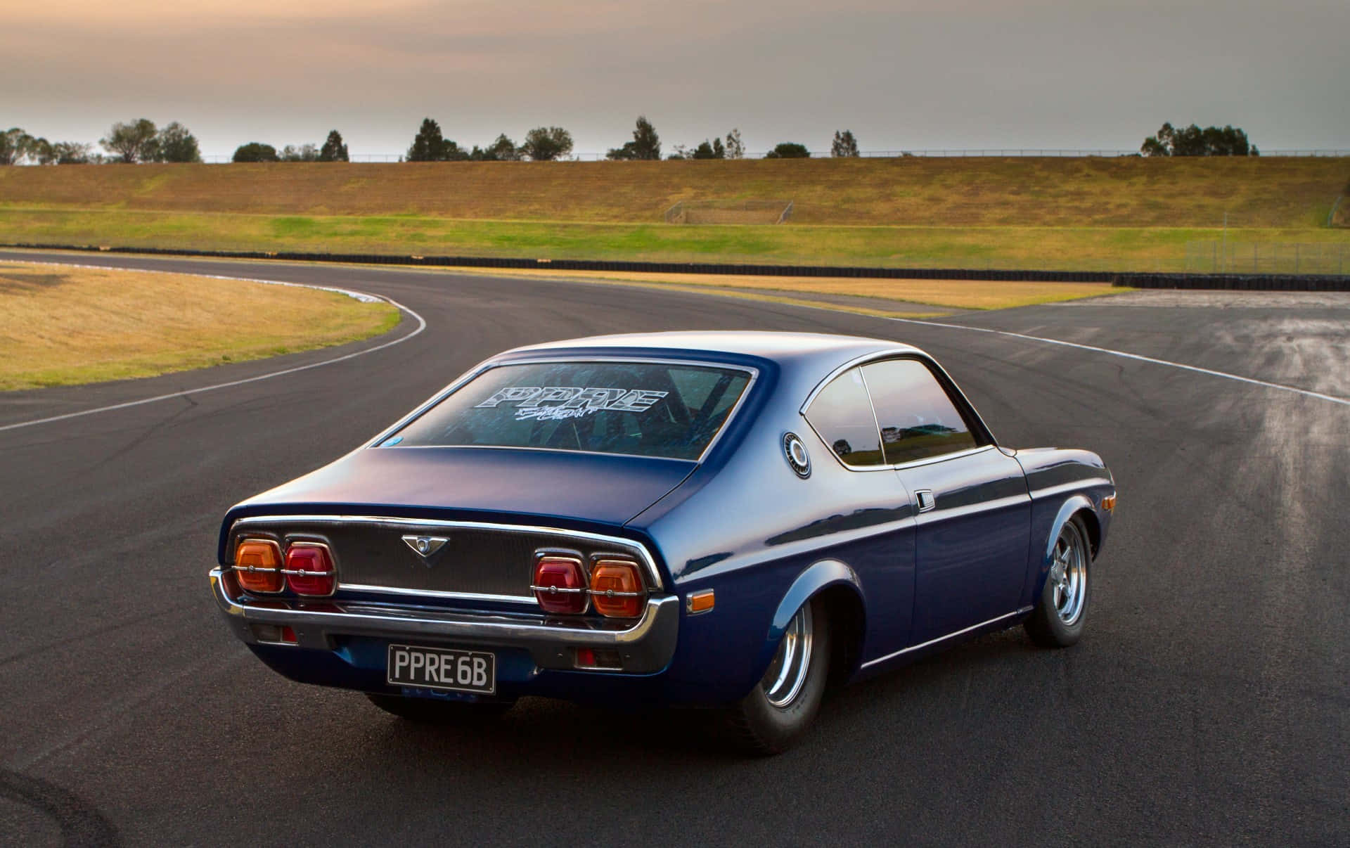 Mazda Rx-3 桌布 壁紙 — 一架流線型藍色經典跑車在蜿蜒的瀝青賽道上行駛，天空呈現柔和的暮色。