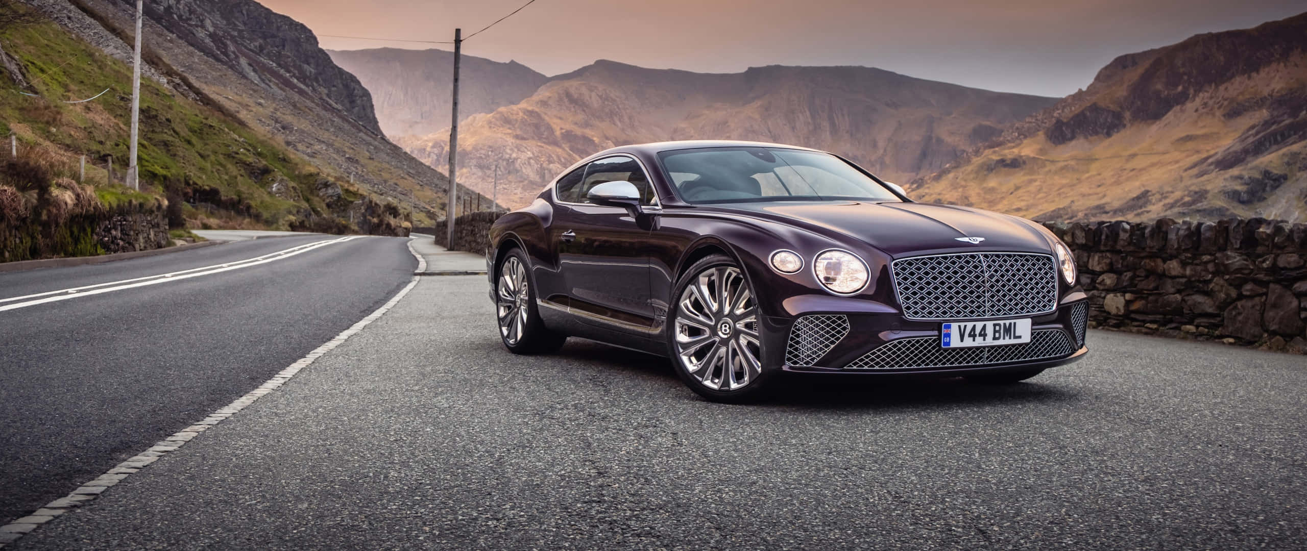 Bentley Mulsanne 桌布 壁紙 — 一架流線型酒紅色賓利 Continental GT 停泊在蜿蜒的山路上，背景是崎嶇的山丘和石牆，天空呈現柔和的金色。