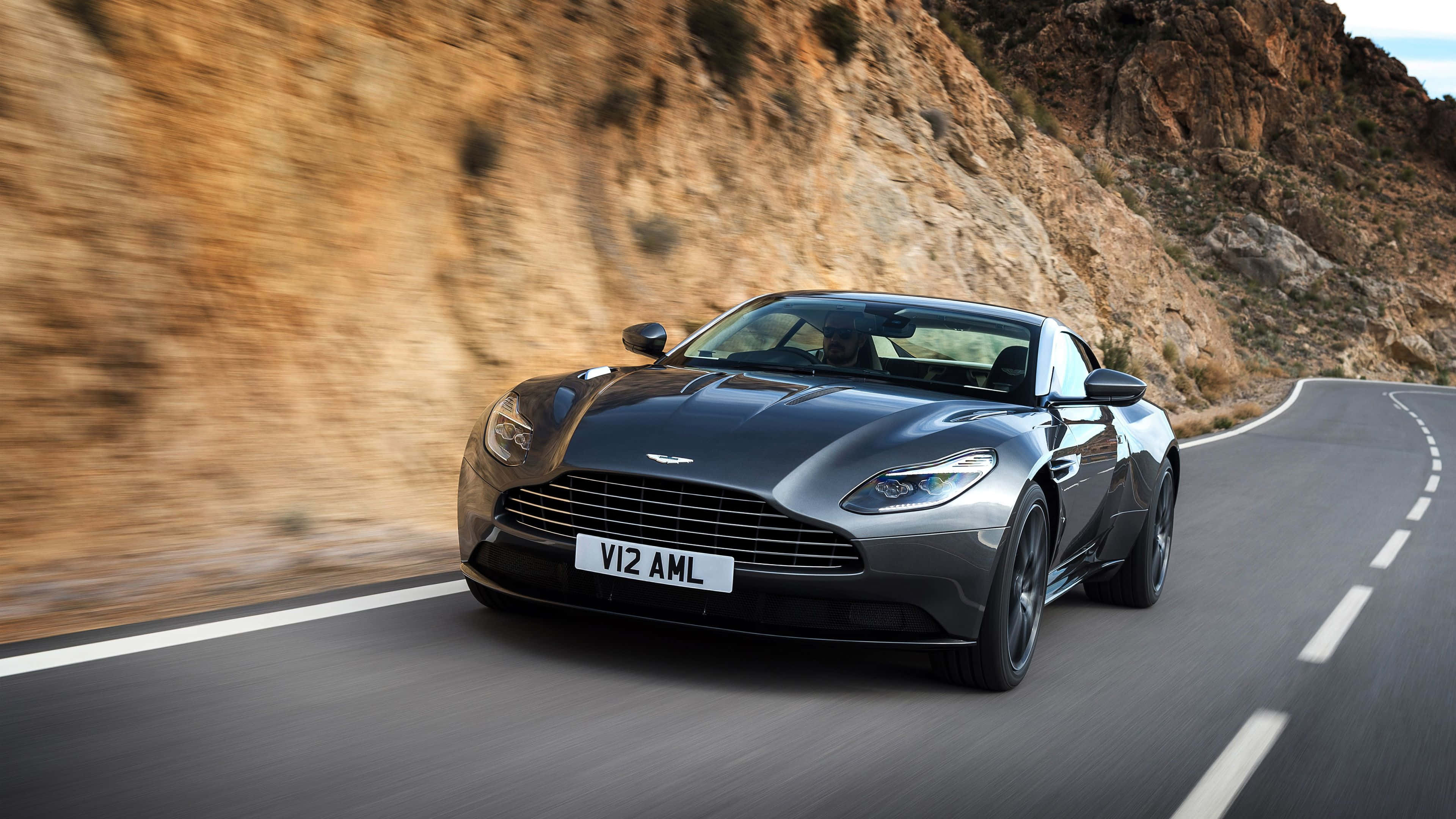 Aston Martin Db5 桌布 壁紙 — 一架流線型銀色 Aston Martin V12 AMI 沿著蜿蜒的山路疾馳，模糊的岩石峭壁呼嘯而過。