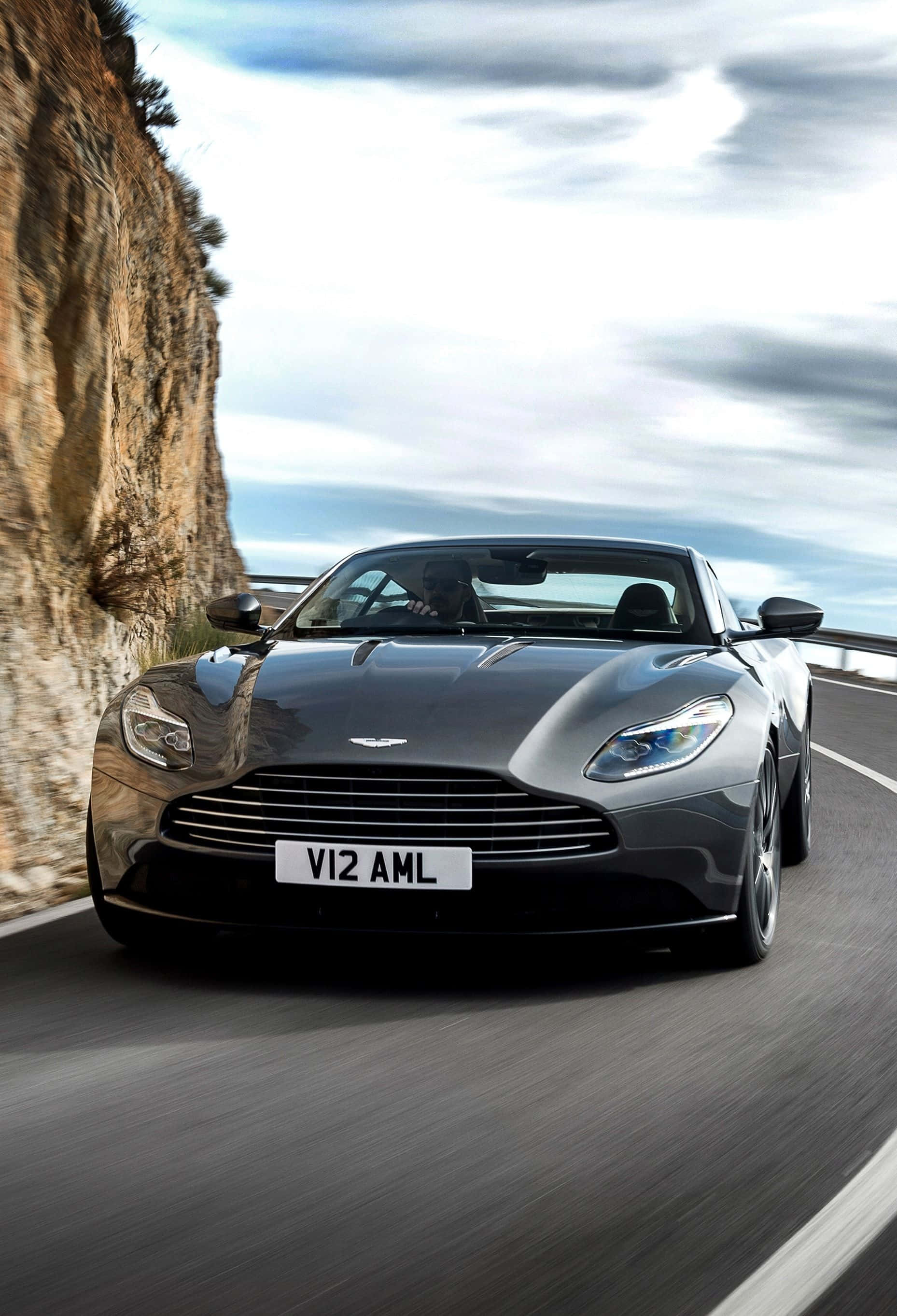 Aston Martin Rapide S 桌布 壁紙 — 一架流線型銀色Aston Martin V12開篷跑車，在多雲的天空下，沿著崎嶇懸崖旁蜿蜒的海岸公路飛馳。