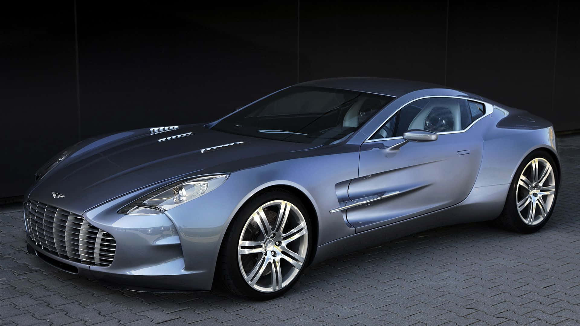 Aston Martin One-77 桌布 壁紙 — 一架流線型銀色Aston Martin One-77跑車在攝影棚燈光下，於深色鋪砌路面上閃閃發光。
