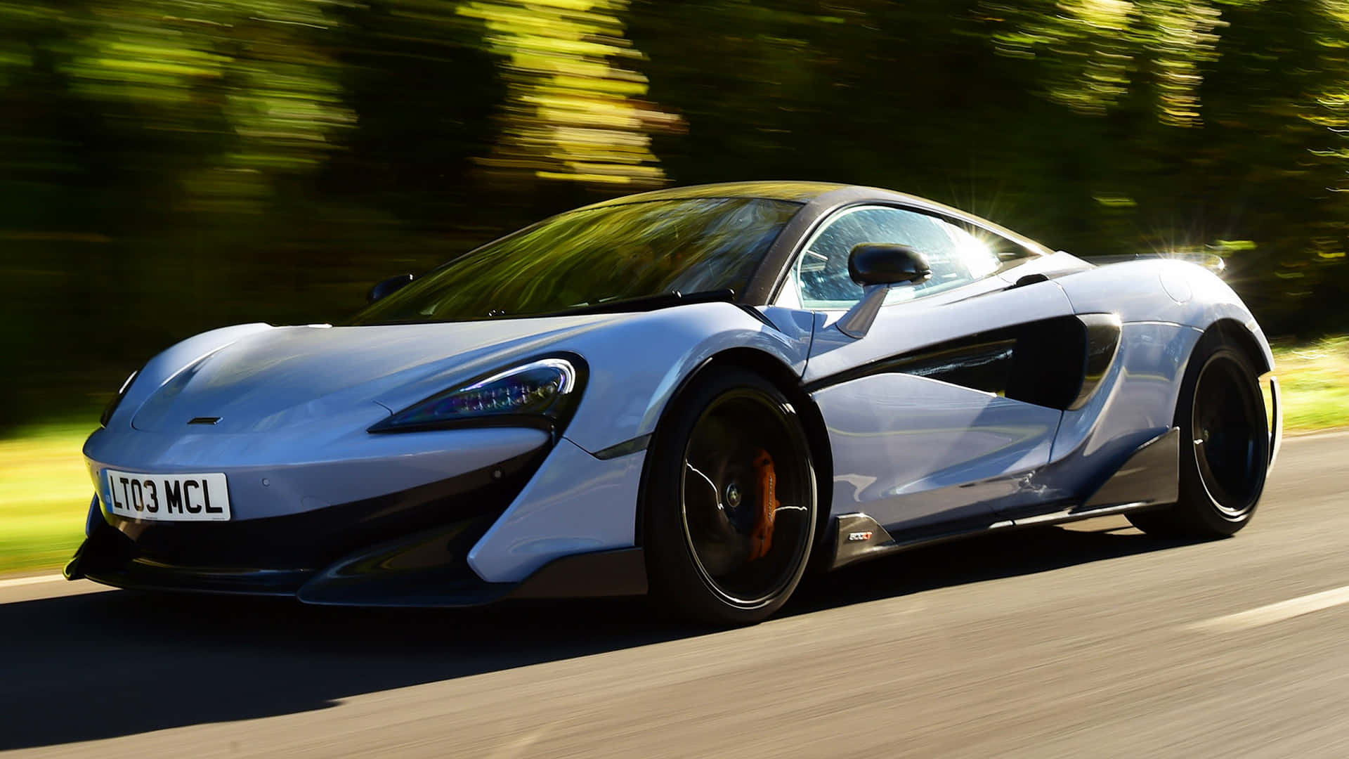 麥拿侖 桌布 壁紙 — 一架流線型銀色McLaren跑車在陽光明媚的道路上行駛，背景是模糊的綠色樹葉。