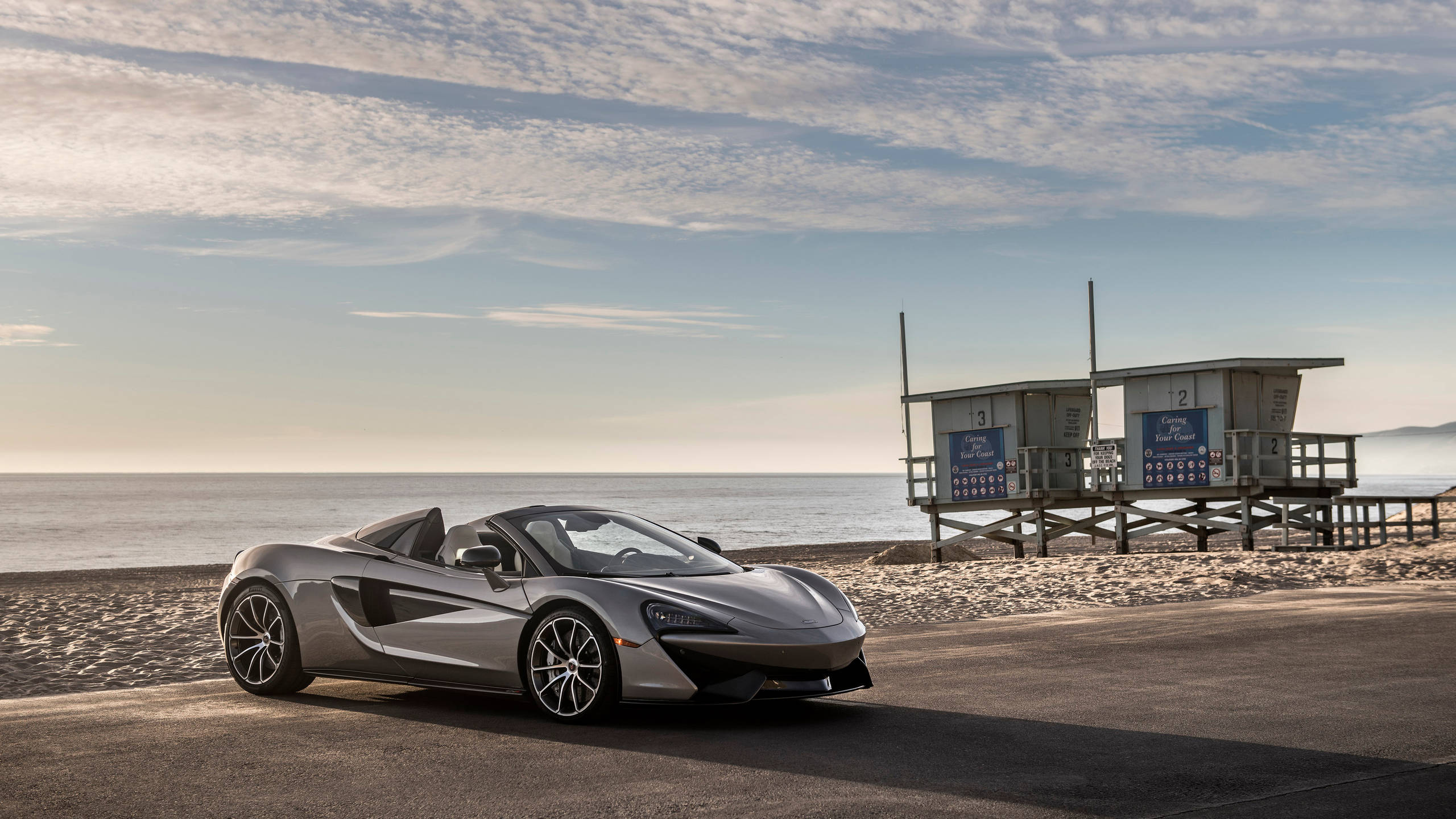 cars 桌布 壁紙 — 一架流線型銀色McLaren跑車停泊在海濱道路上，旁邊有救生員瞭望塔，背景是海洋和天空。