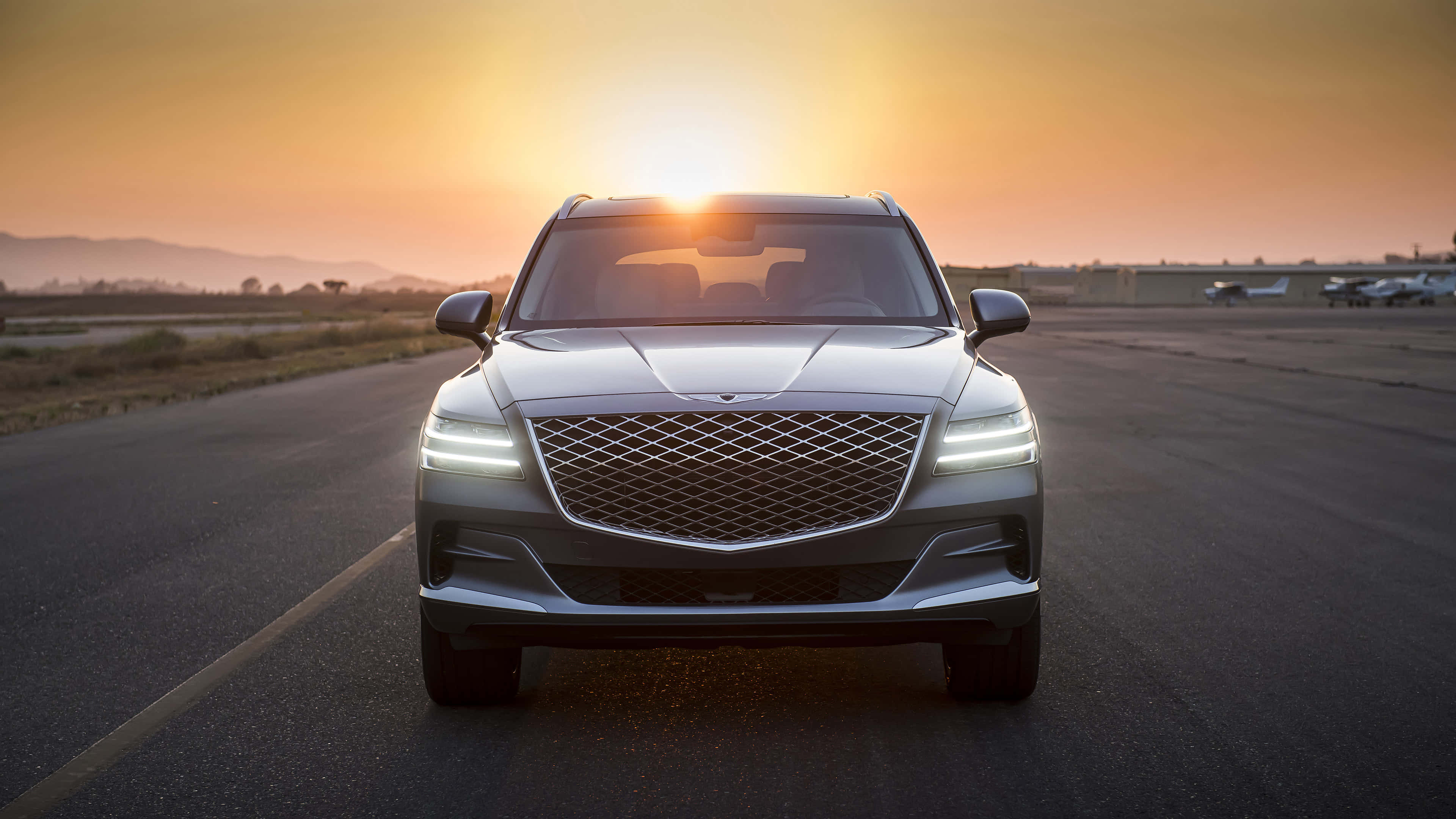 Genesis Gv80 桌布 壁紙 — 一架流線型銀色SUV在空曠的跑道上向前行駛，夕陽在其後方落下，散發出溫暖的金色光芒。