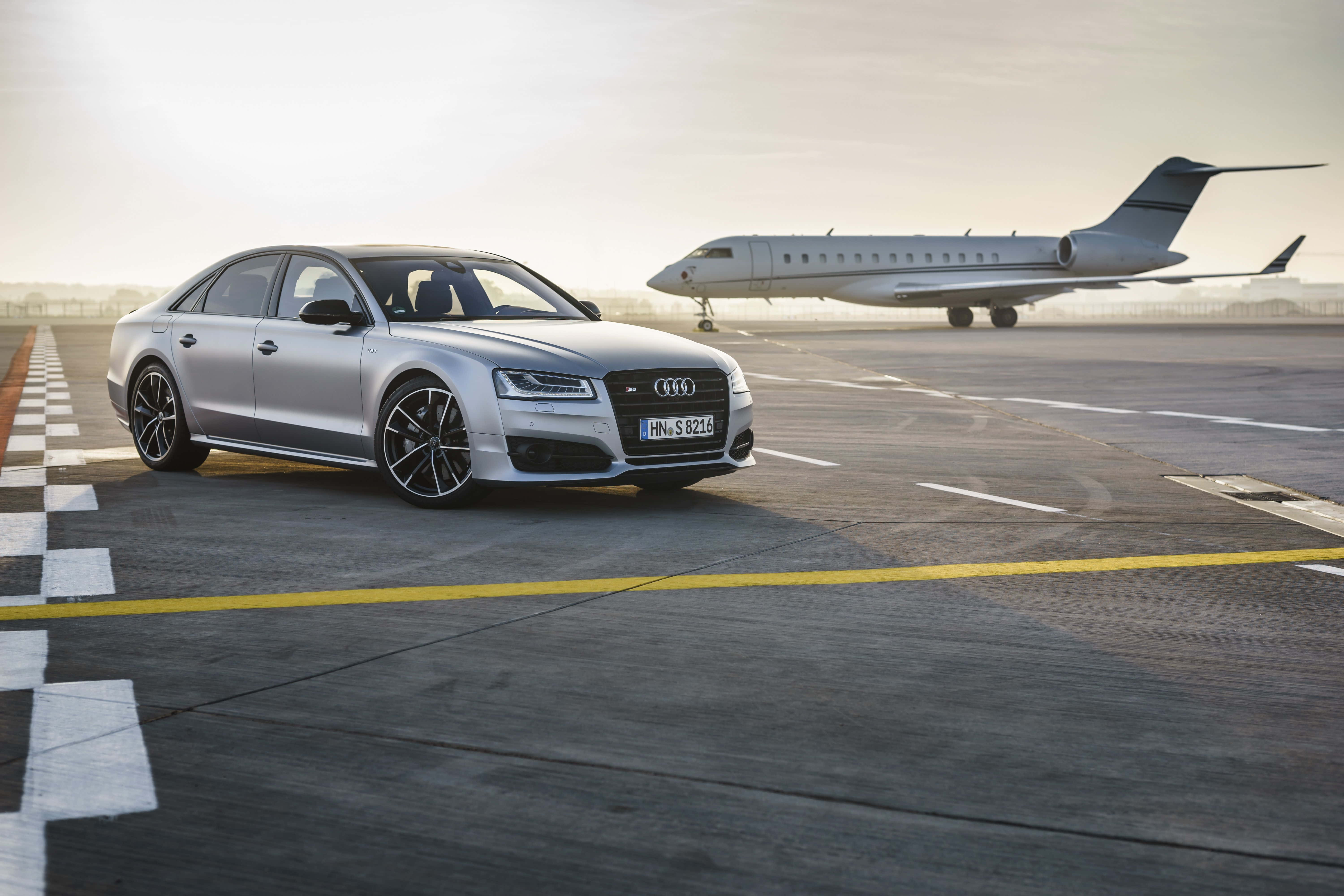 Audi S8 桌布 壁紙 — 一架流線型銀色奧迪S8房車停在機場停機坪上，背景有私人飛機，沐浴在柔和的金色光線中。