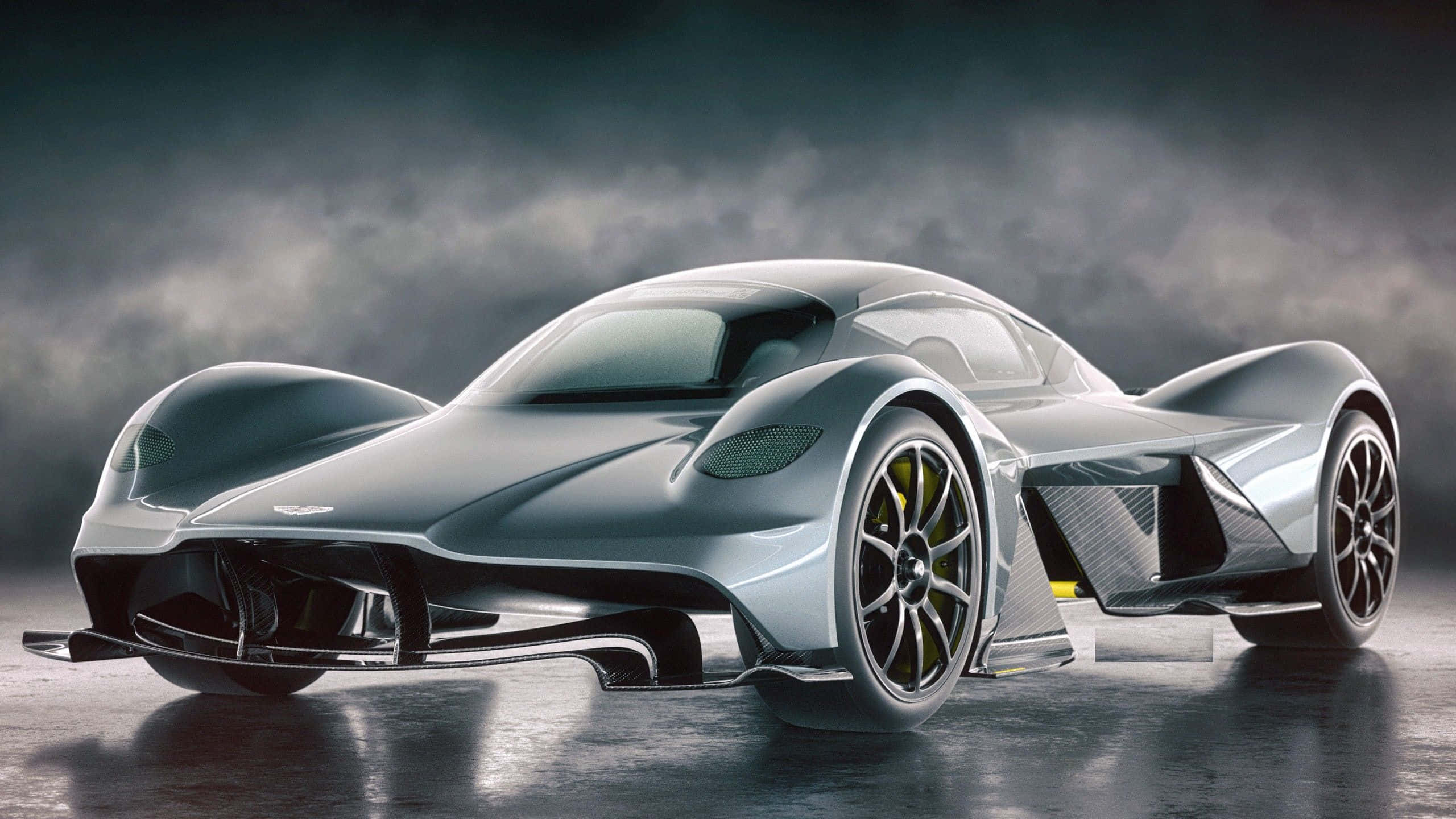 Aston Martin Valkyrie 桌布 壁紙 — 一架流線型銀色未來感跑車，配以發光黃色制動卡鉗，在戲劇性的暴風雨天空下，立於深色反光表面上。