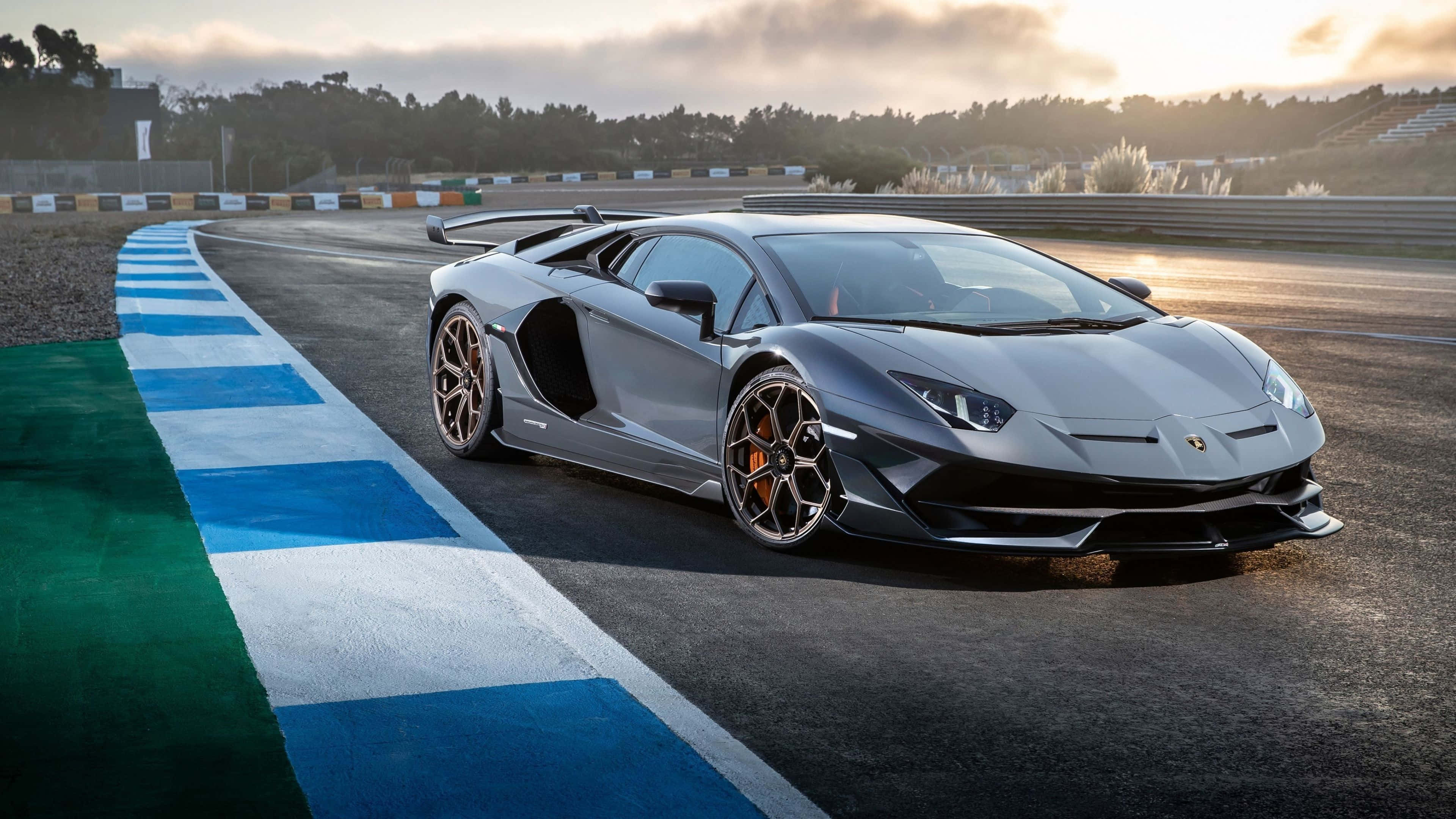 林寶堅尼 4K 桌布 壁紙 — 一架流線型銀色林寶堅尼 Aventador SVJ 在陽光明媚的賽道上奔馳，賽道有藍白路肩，遠處有看台，天空多雲。