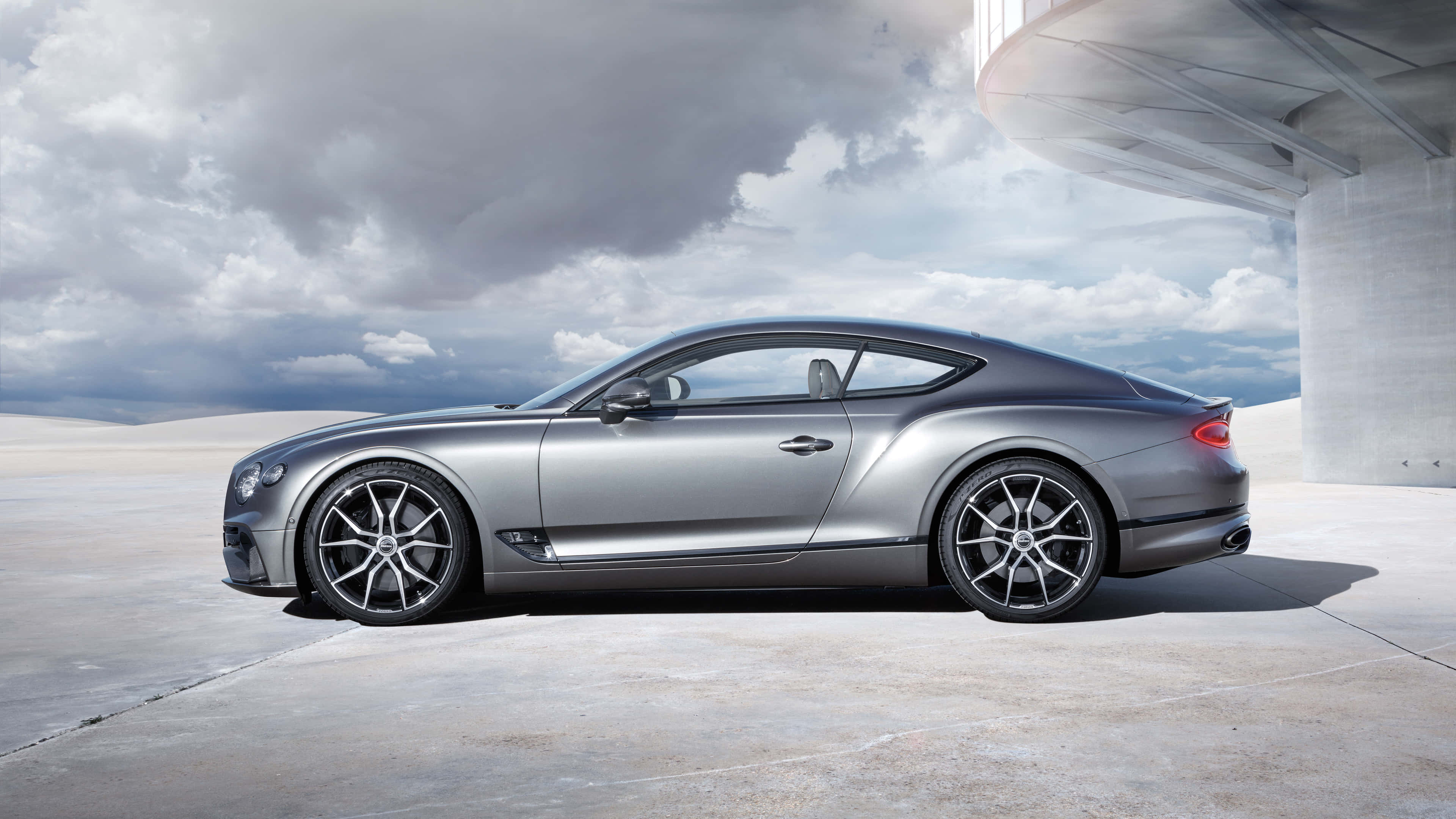 Bentley Continental Gt 桌布 壁紙 — 一架流線型銀色賓利 Continental GT 雙門跑車停泊在現代混凝土結構下，背景為多雲的天空。