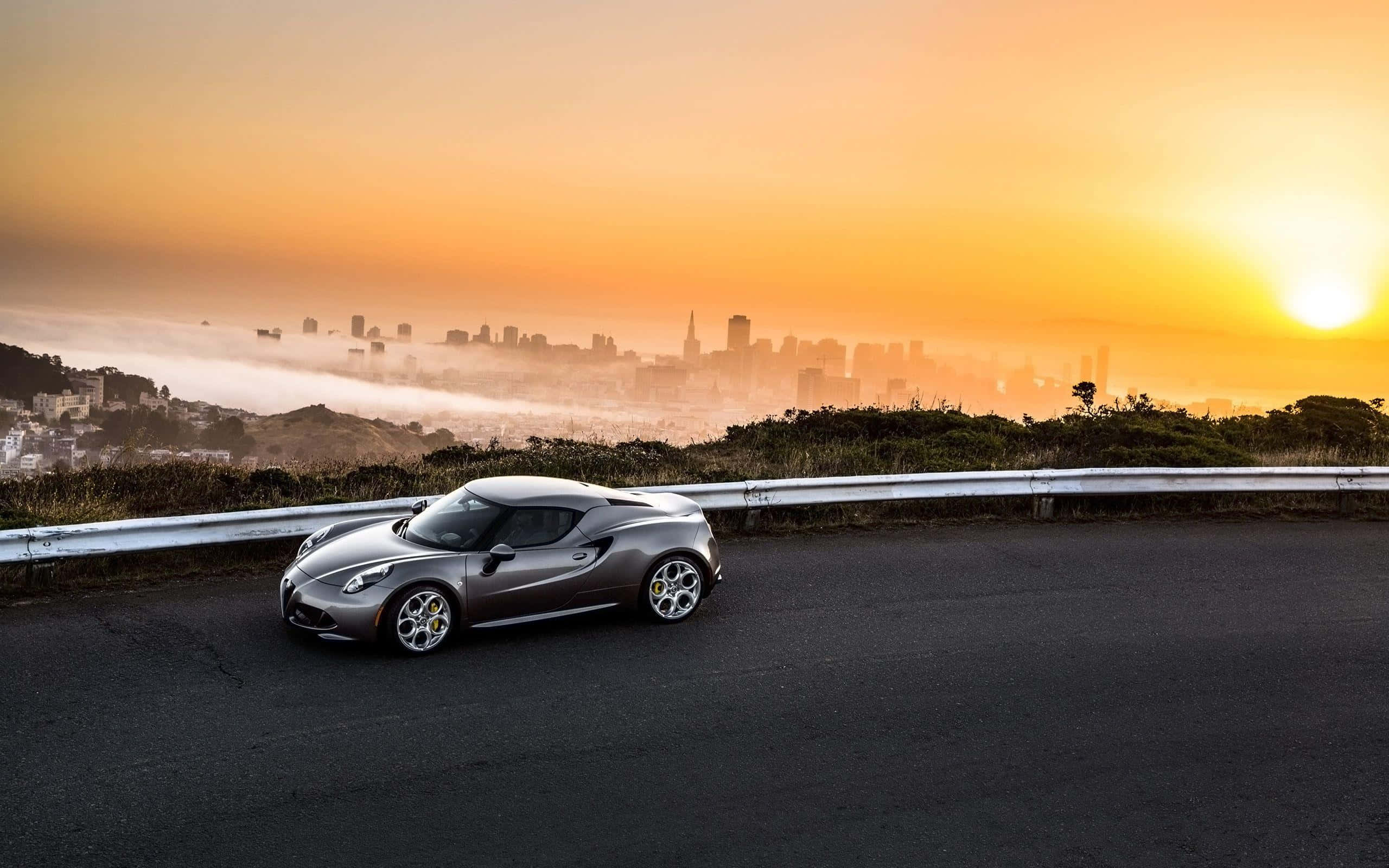 Alfa Romeo 4c 桌布 壁紙 — 一架流線型銀色跑車停泊在蜿蜒道路上，俯瞰著金色日落時分霧氣朦朧的城市天際線。