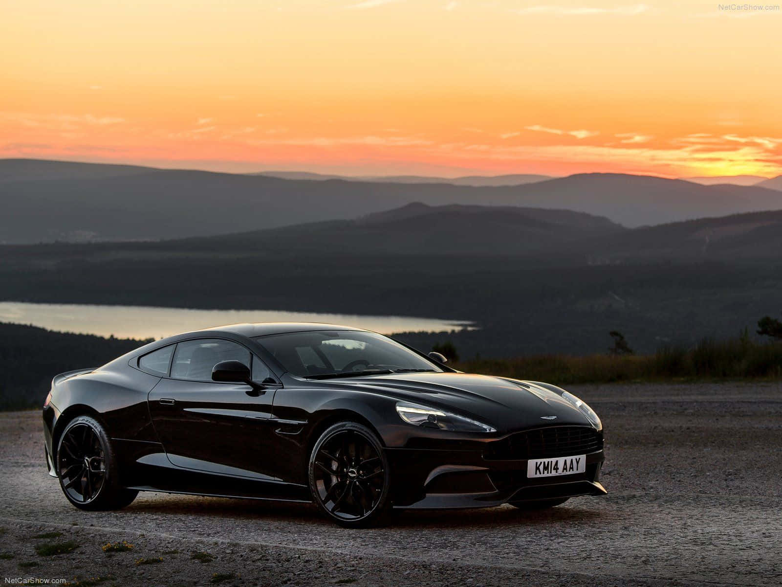 Aston Martin Vanquish 桌布 壁紙 — 一架流線型黑色 Aston Martin V12 Vantage 停泊在日落時分的碎石觀景點，背景是連綿起伏的山丘和湖泊。