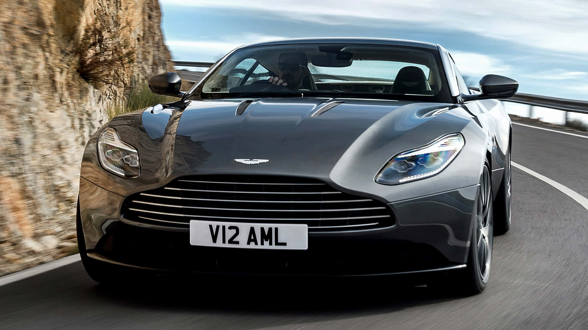 Aston Martin Db11 桌布 壁紙 — 一架流線型黑色Aston Martin跑車沿著蜿蜒的山路疾馳，旁邊是岩石峭壁。