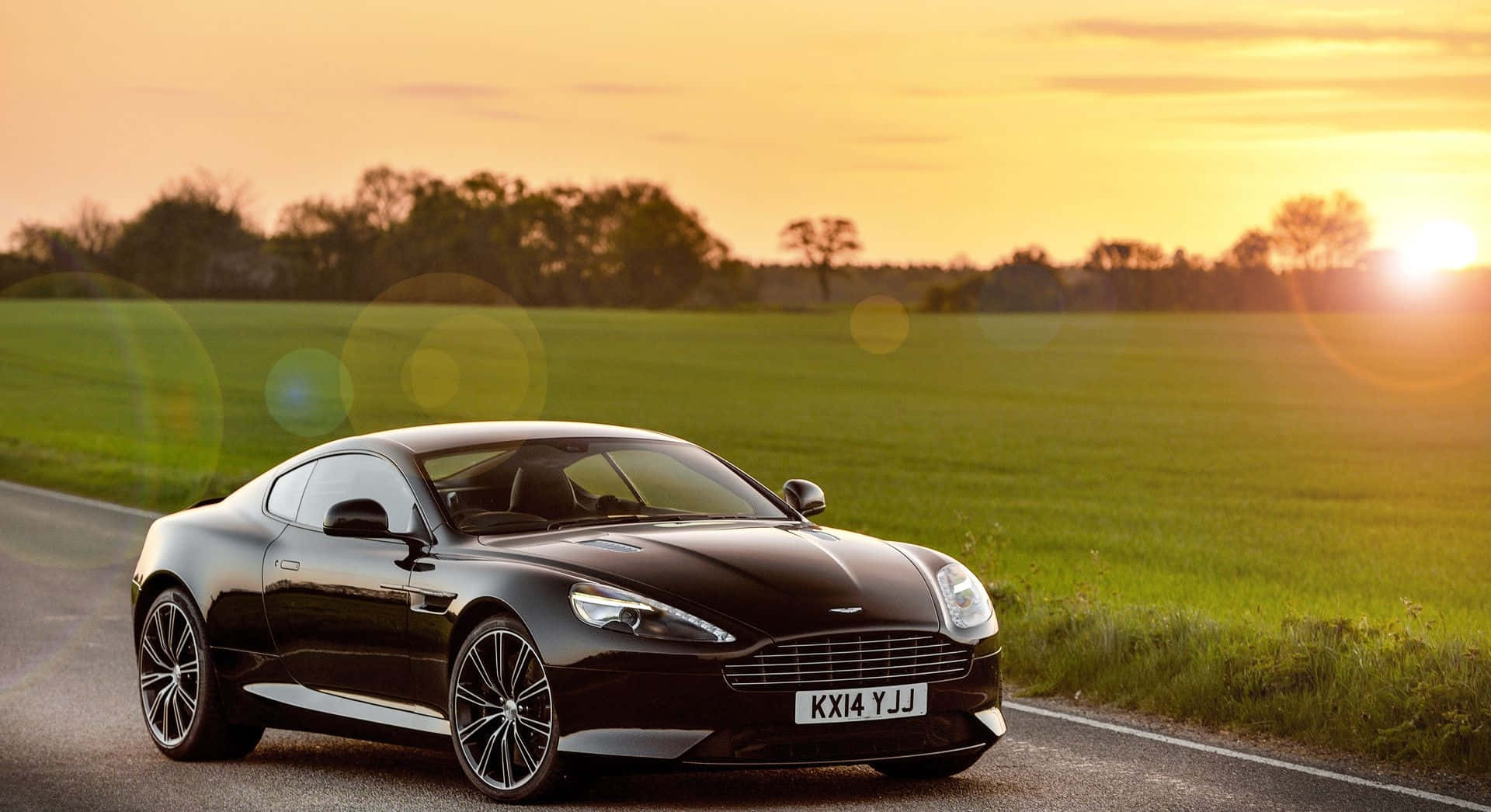 Aston Martin Db9 桌布 壁紙 — 一架流線型黑色Aston Martin跑車在金色夕陽與鏡頭光暈中沿鄉村道路滑行。