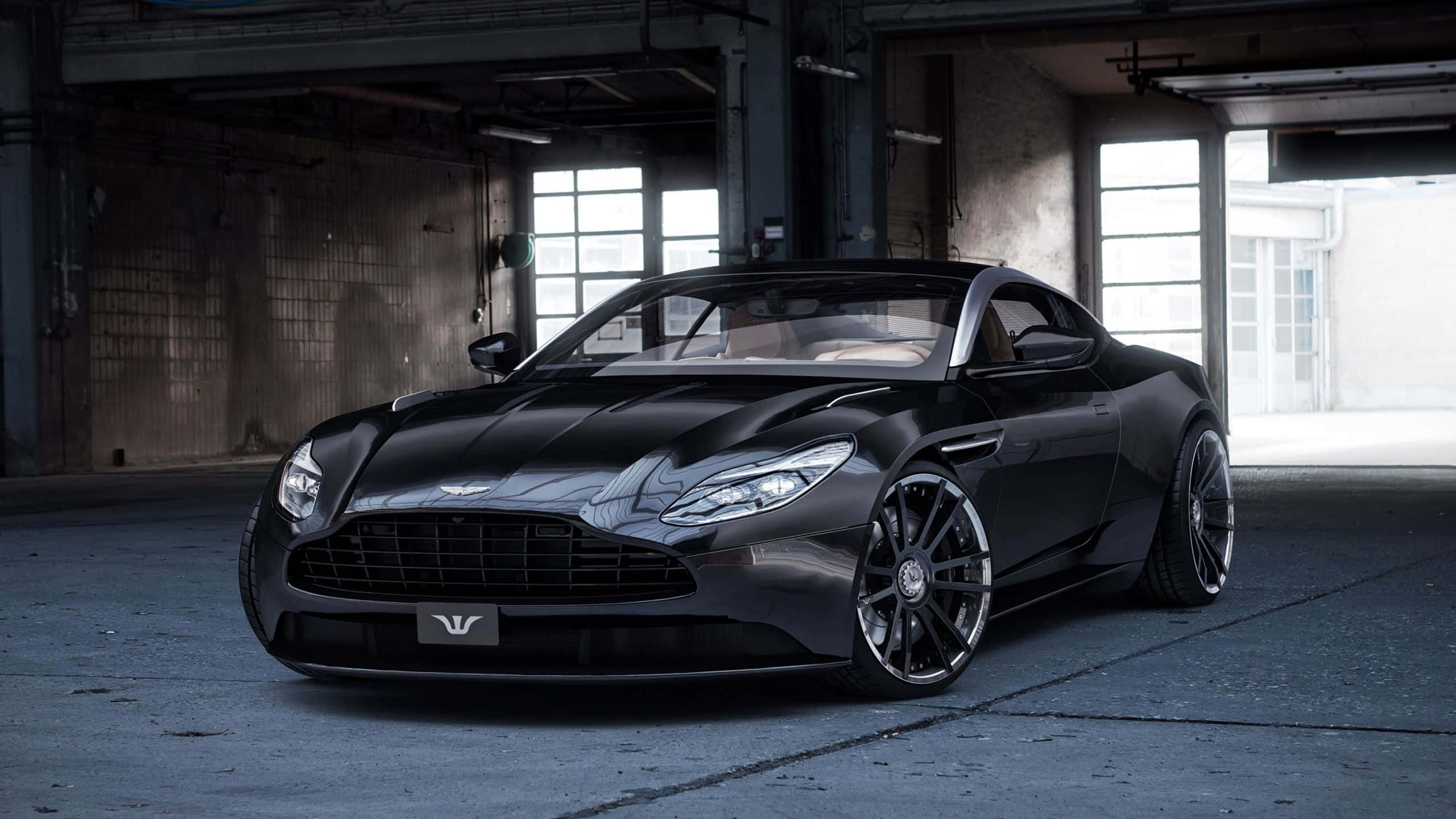 Aston Martin Vanquish 桌布 壁紙 — 一架流線型黑色Aston Martin跑車在工業照明下，於粗糙的倉庫內閃閃發光。
