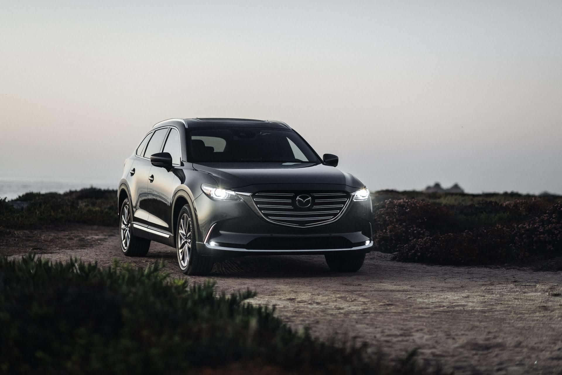 Mazda Cx-9 桌布 壁紙 — 一架流線型黑色Mazda CX-9 SUV停泊在海岸泥路上，車頭燈亮起，映襯著柔和嘅暮色天空。