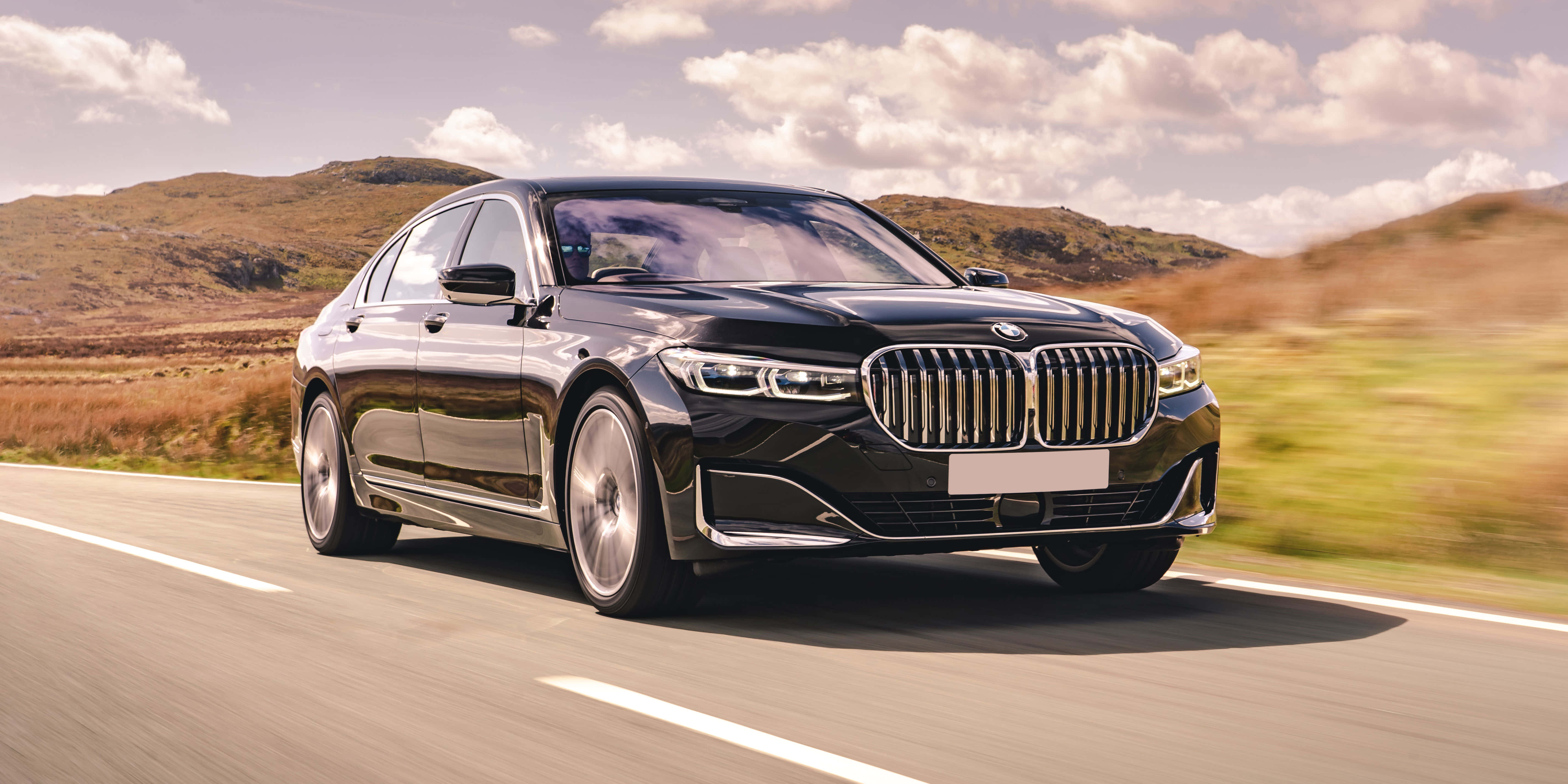 Bmw 7 Series 桌布 壁紙 — 一架流線型黑色寶馬房車在部分多雲的天空下，沿著蜿蜒的道路穿過起伏的金色山丘疾馳。