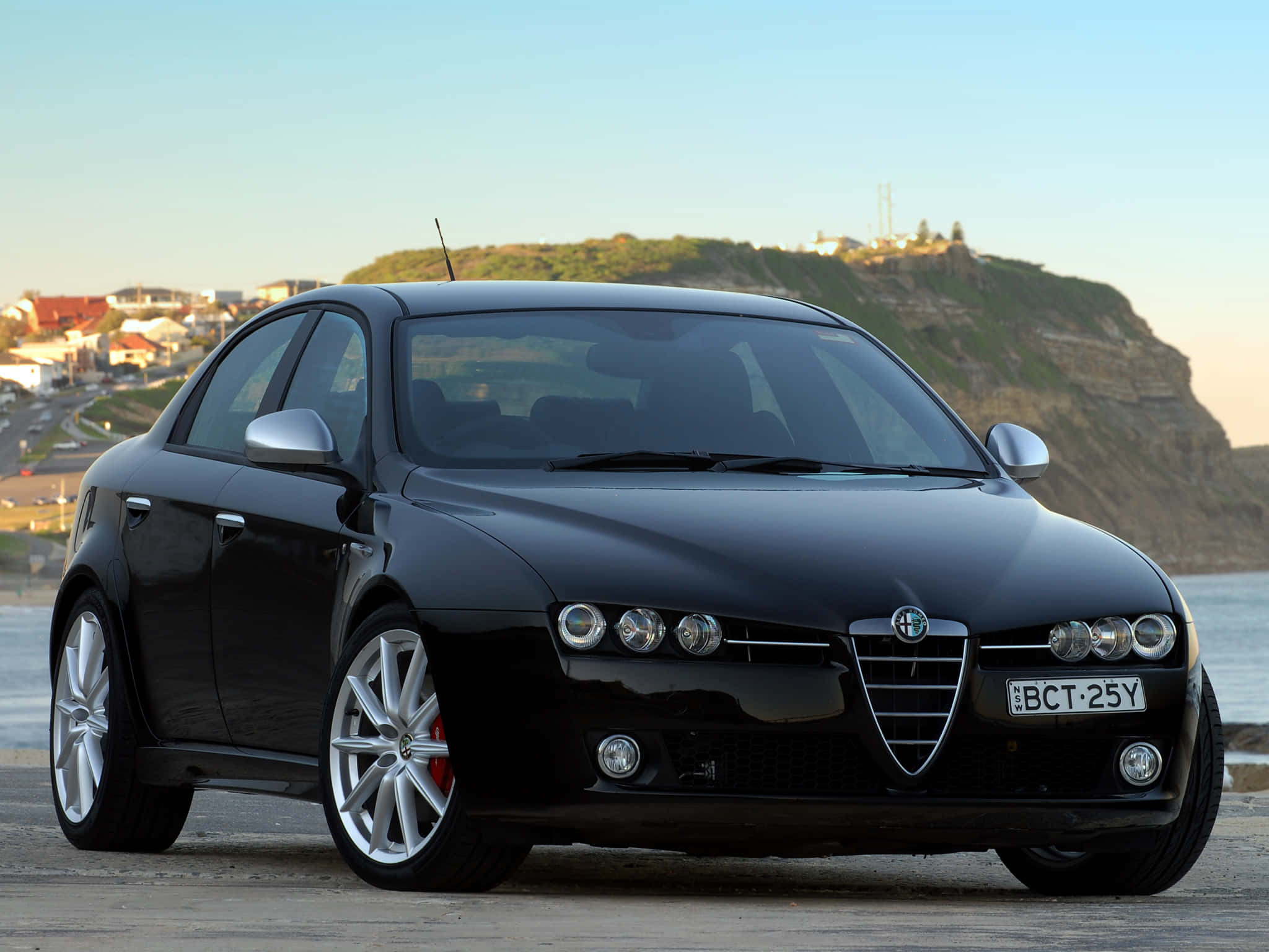 Alfa Romeo 159 桌布 壁紙 — 一架流線型黑色愛快羅密歐房車停泊在沿海公路上，背景是懸崖小鎮和海洋。