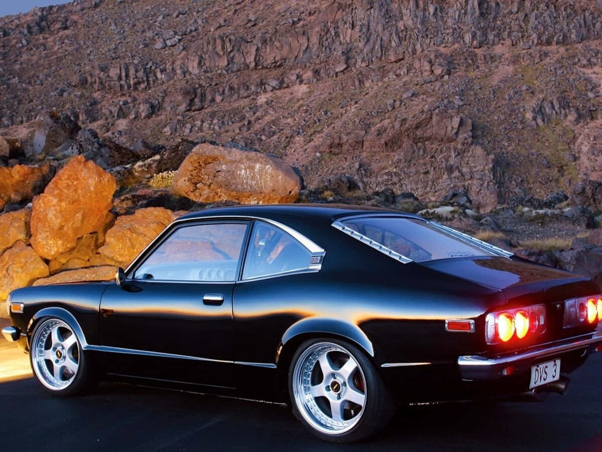 Mazda Rx-3 桌布 壁紙 — 一架流線型黑色經典跑車，帶有發光的紅色尾燈，停在沙漠公路上，旁邊是崎嶇的橙色和棕色岩層。