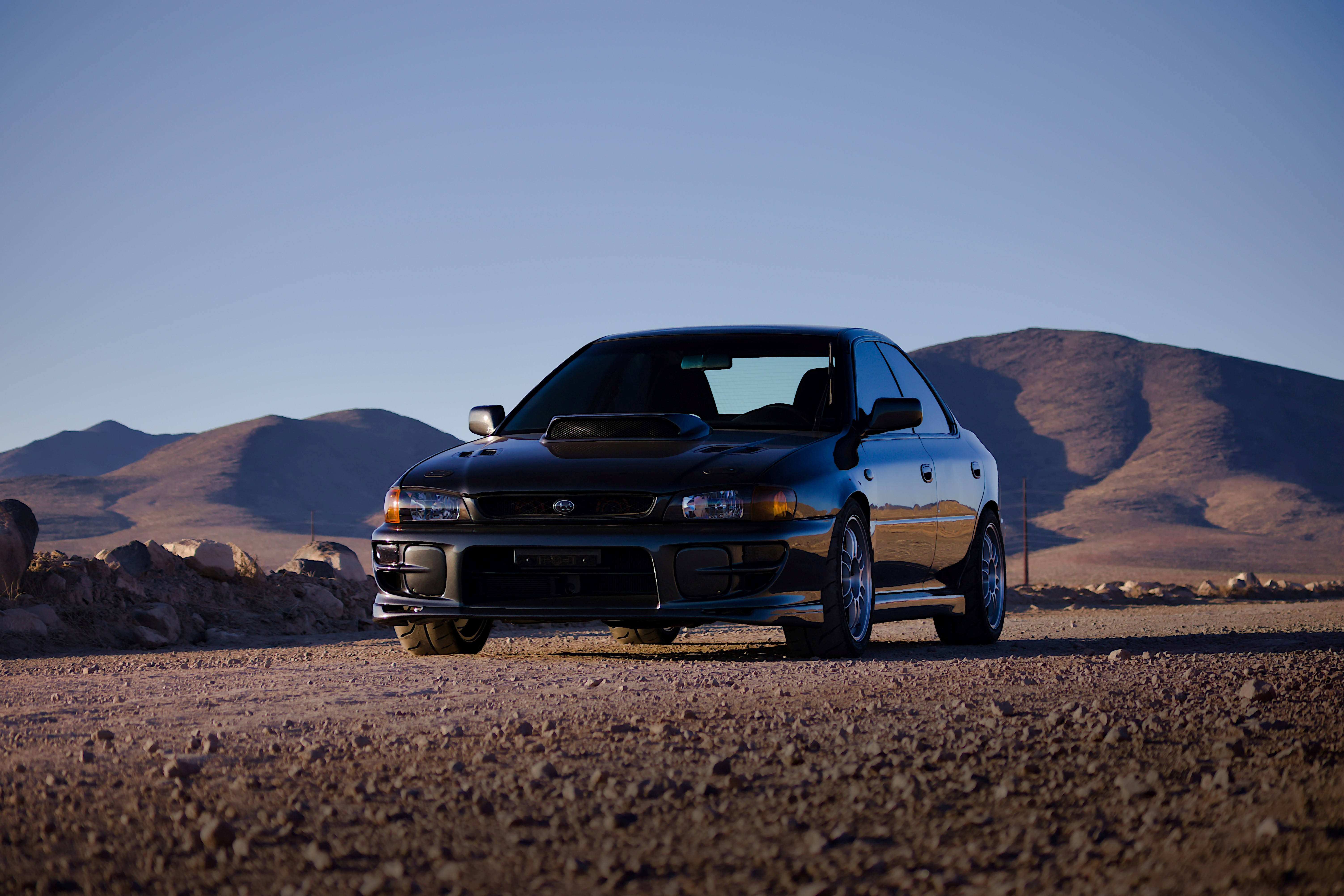 Dirt Rally Subaru Impreza Turbo GC 壁紙 — 一架流線型黑色跑車停泊在岩石沙漠道路上，遠山在晴朗藍天下。