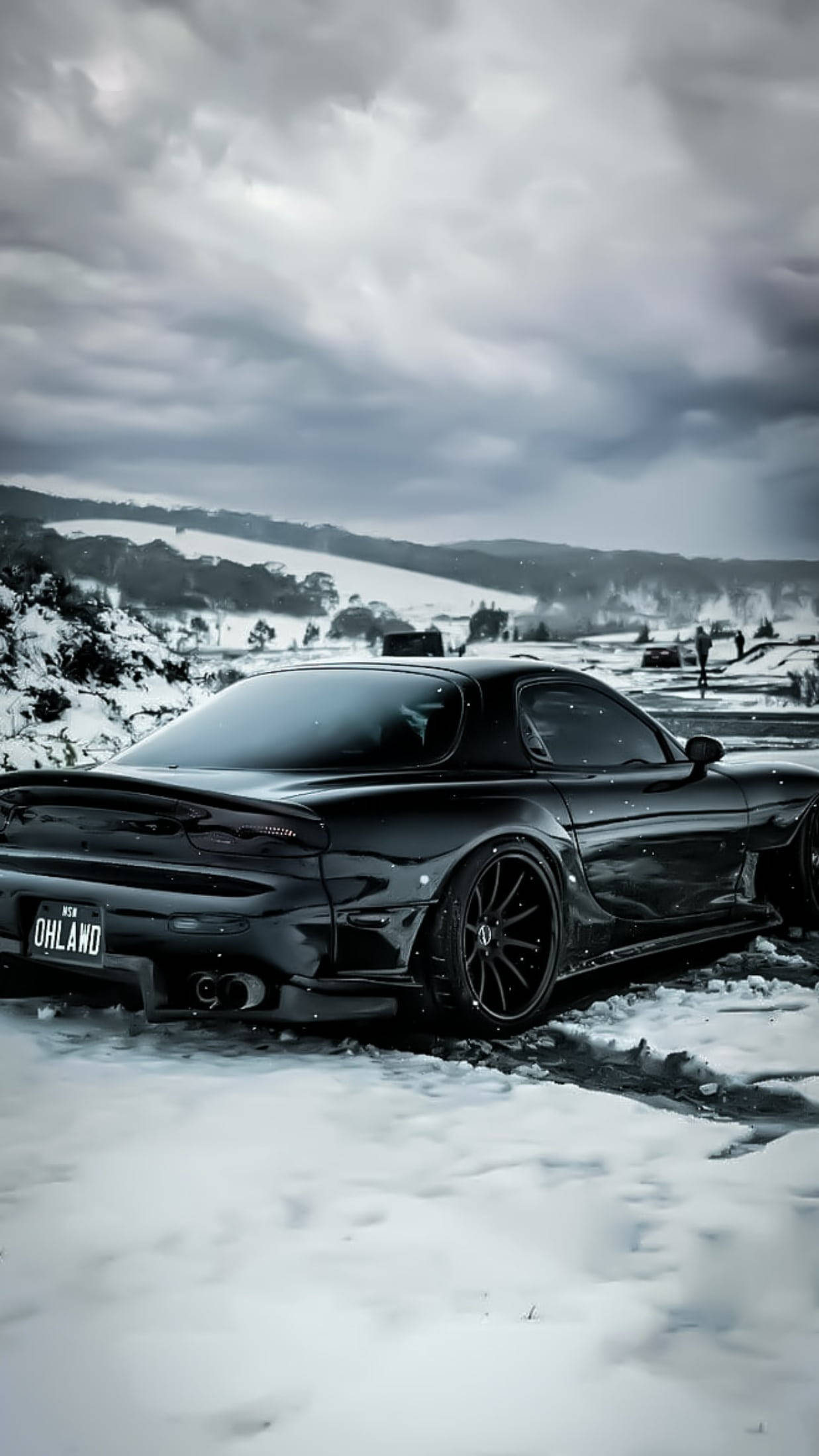 Mazda 桌布 壁紙 — 一架流線型黑色跑車停泊在雪地上，背景是戲劇性的多雲天空和連綿起伏的山丘。