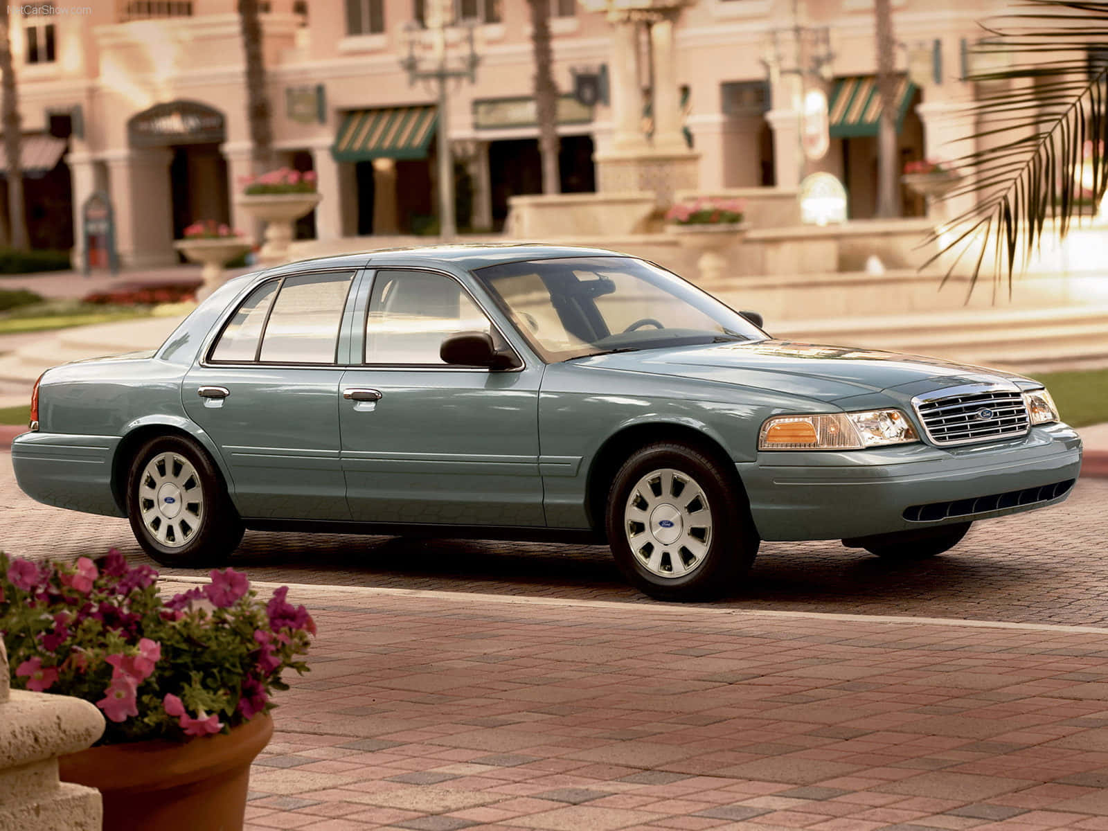 Ford Crown Victoria 桌布 壁紙 — 一架淺藍色福特皇冠維多利亞轎車停泊在磚砌廣場上，周圍有棕櫚樹和商店門面，沐浴在溫暖的黃金時刻光線中。