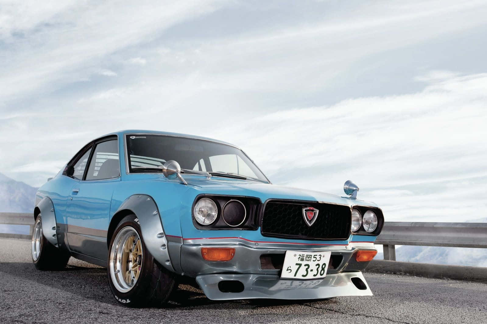 Mazda Rx-3 桌布 壁紙 — 一架淺藍色經典日本跑車，配備寬體套件和金色輪圈，停泊在陰天天空下的山路上。