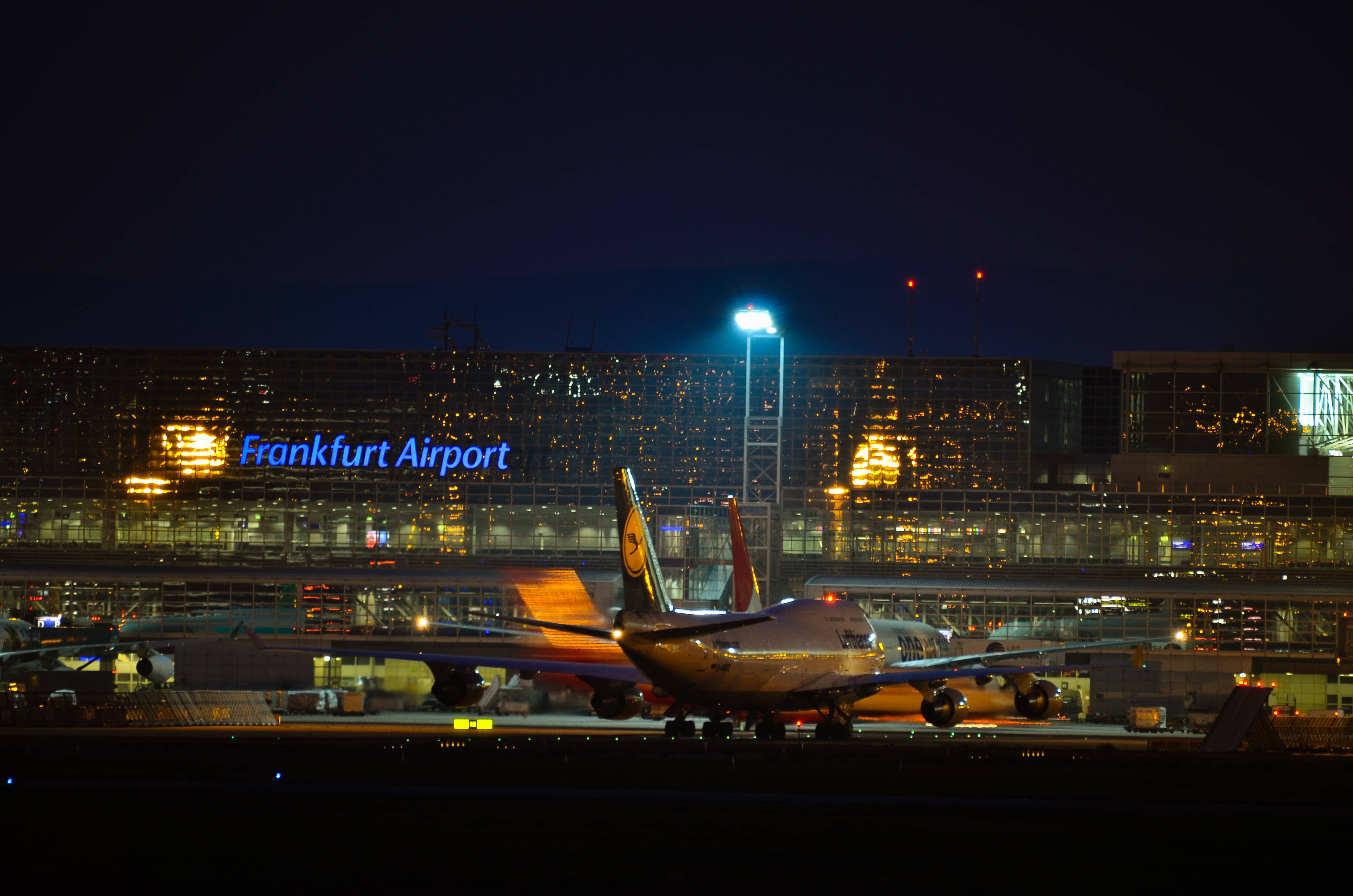 Airport 桌布 壁紙 — 一架漢莎航空飛機停在法蘭克福機場停機坪上，在黑暗的天空下被航站樓燈光和明亮的塔燈照亮。