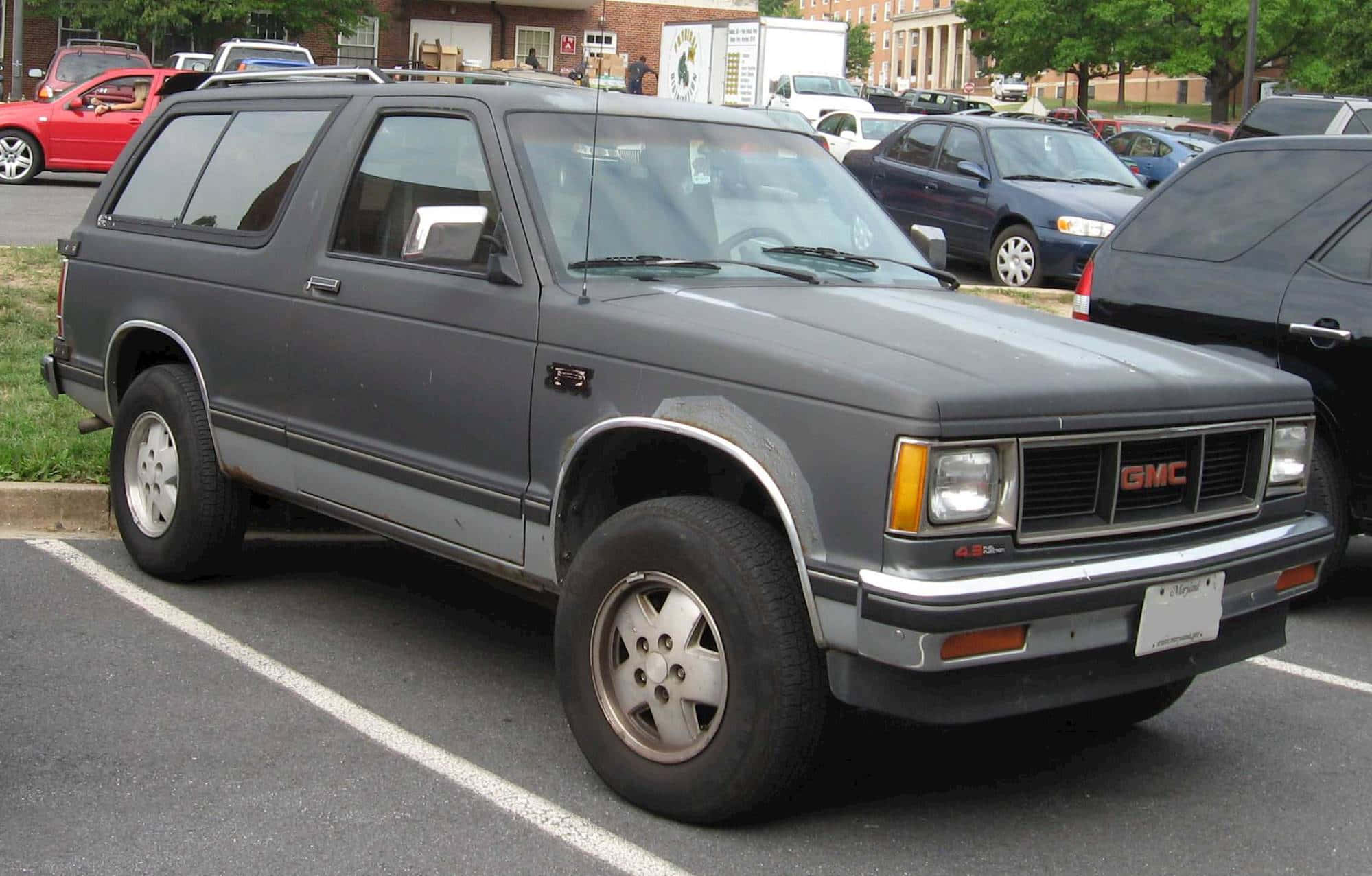 Gmc Jimmy 桌布 壁紙 — 一架灰色1980年代GMC Safari SUV停泊在日間停車場，背景有其他汽車和磚砌建築。