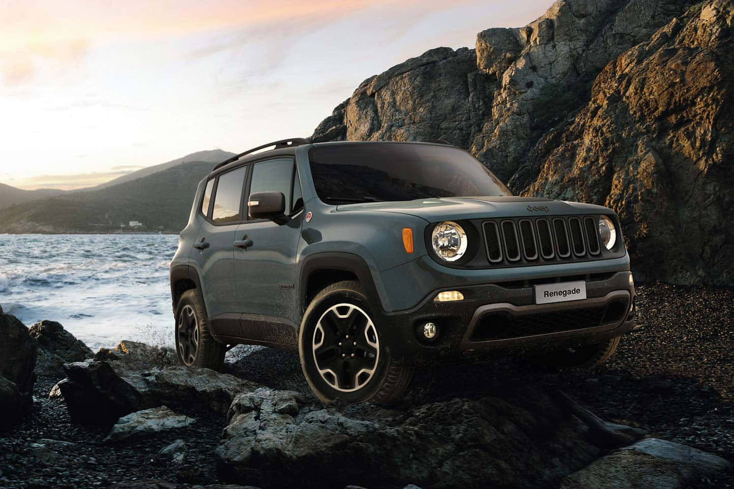 Jeep Renegade 桌布 壁紙 — 一架灰色Jeep Renegade SUV停泊在岩石地形上，旁邊是日落時分波濤洶湧的海洋，背景有山脈。