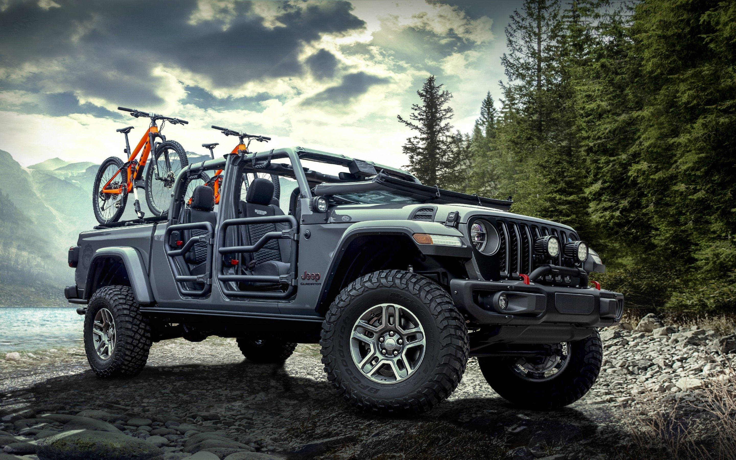 Jeep Gladiator Rubicon 壁紙 — 一架灰色Jeep Wrangler車頂綁著兩輛橙色山地自行車，停在岩石湖岸上，天空多雲而戲劇化。