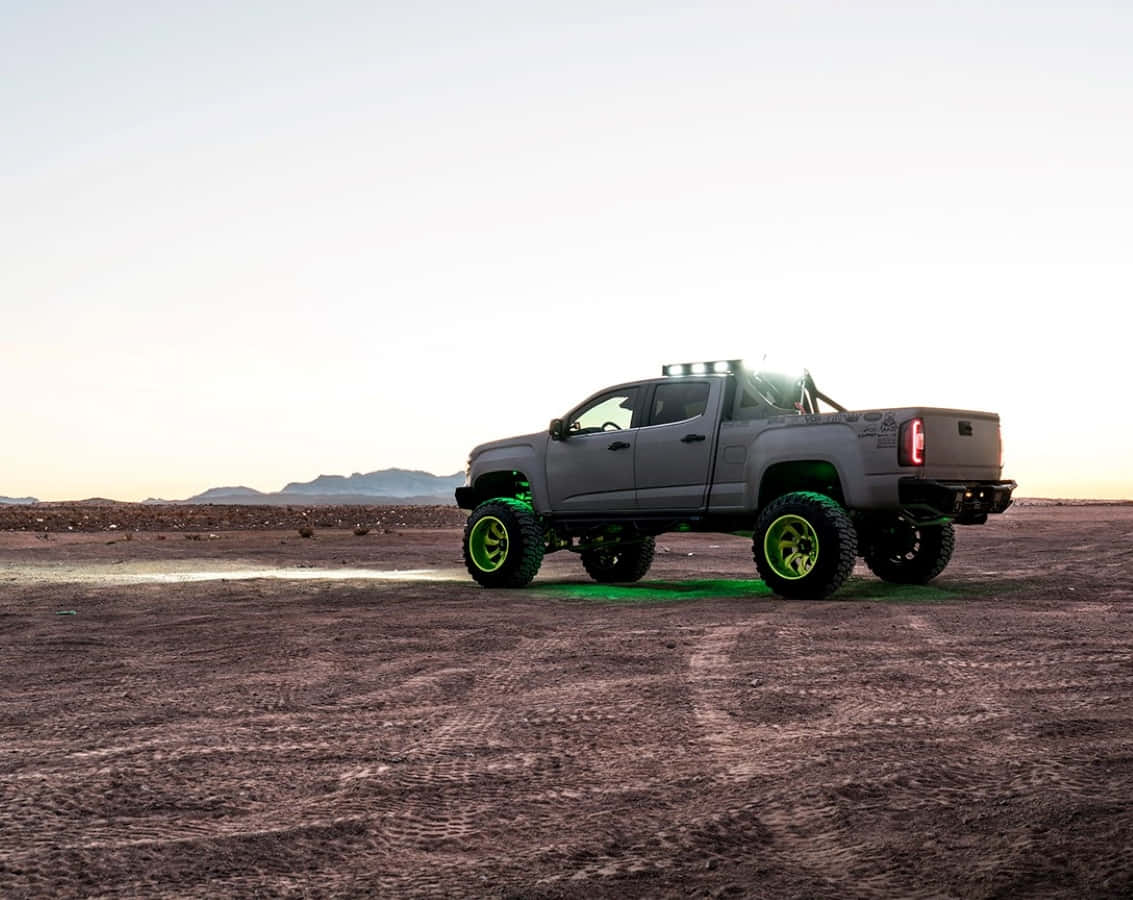 Gmc Canyon 桌布 壁紙 — 一架灰色升高嘅皮卡車，帶有發光綠色車輪，企喺廣闊嘅沙漠中，喺日落時分喺沙地上投下長長嘅影子。