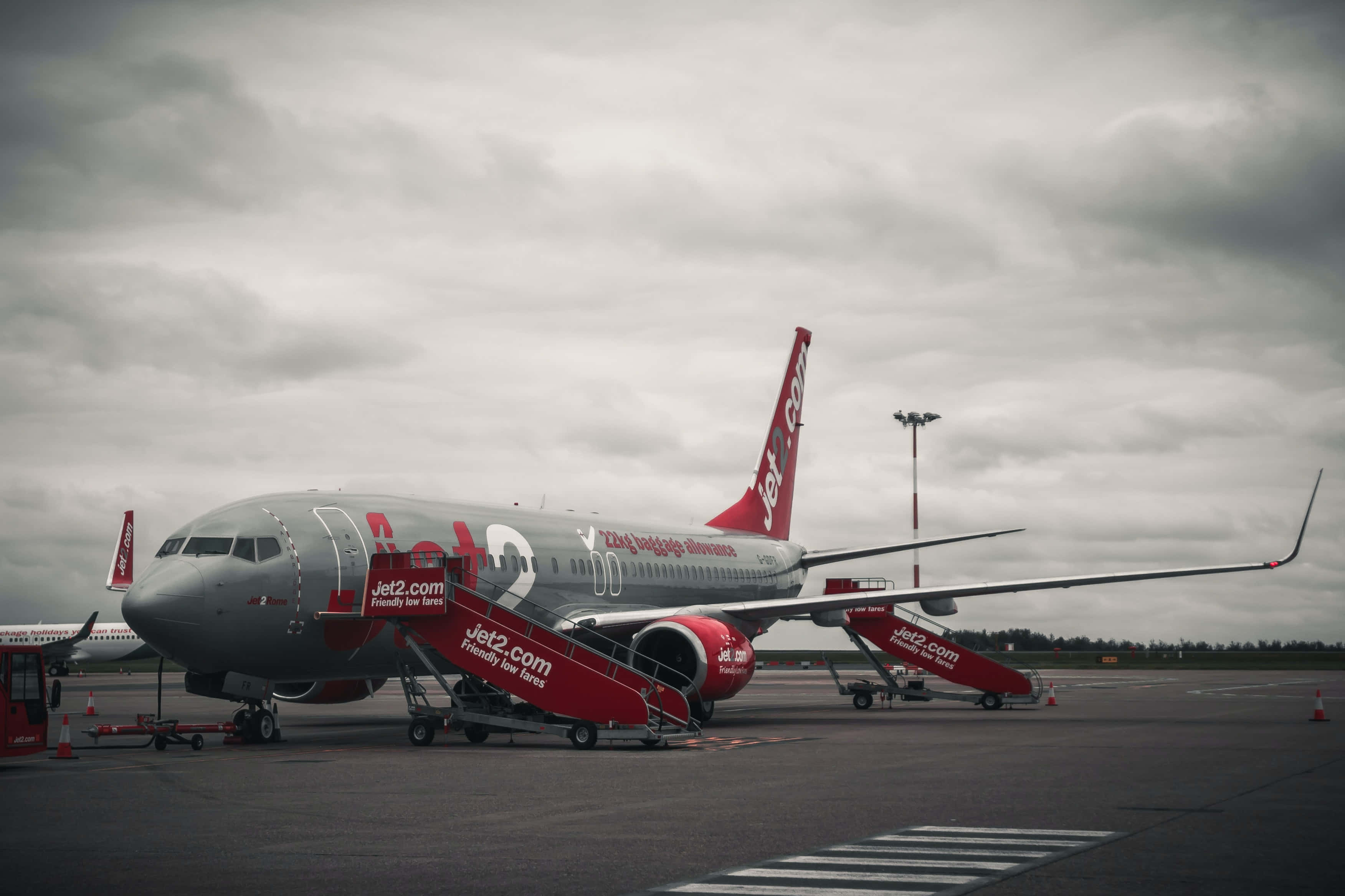 Airport 桌布 壁紙 — 一架灰色和紅色的Jet2.com飛機停在潮濕的停機坪上，天空多雲，並連接了登機梯。