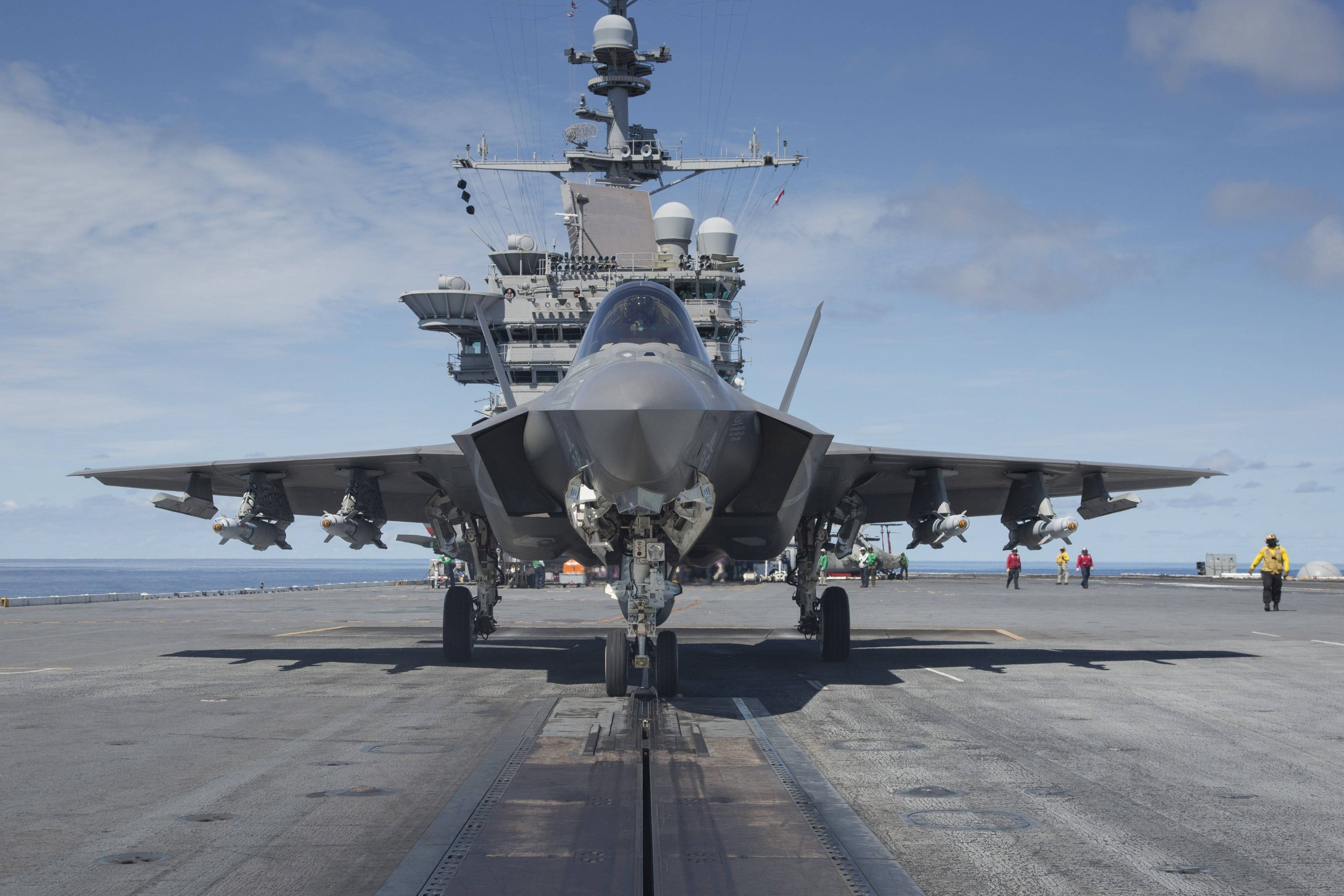 飛機 桌布 壁紙 — 一架灰色戰鬥機停放在航空母艦甲板上，背景是艦島和海洋，天空局部多雲。