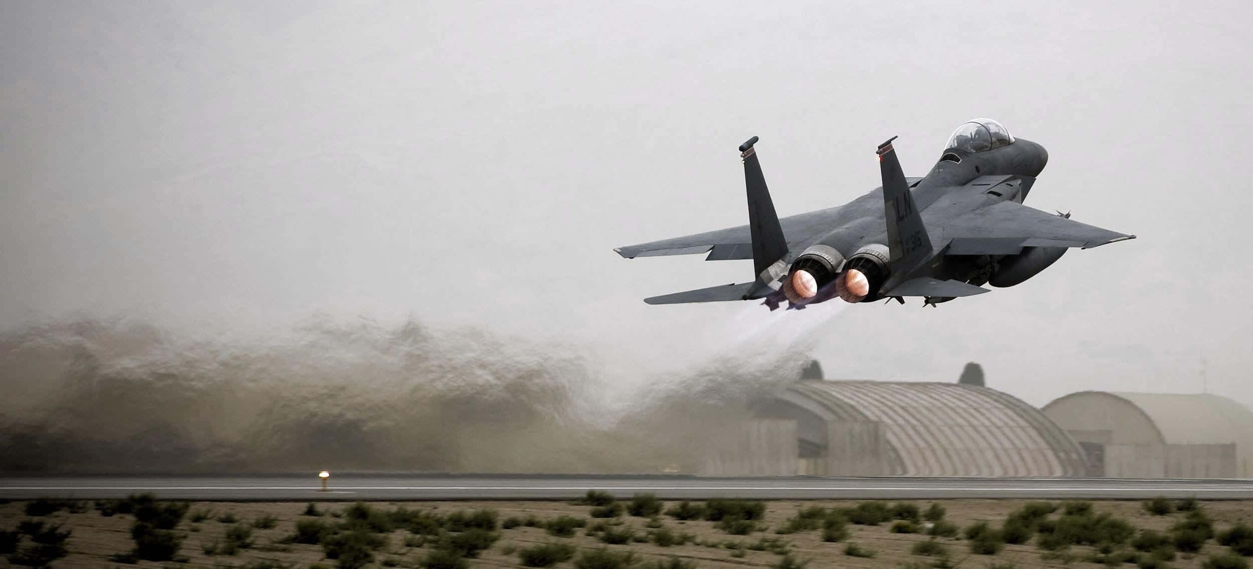 Air Force 桌布 壁紙 — 一架灰色戰鬥機從沙漠跑道起飛，揚起塵土，加力燃燒室發出光芒，背景是朦朧的天空。