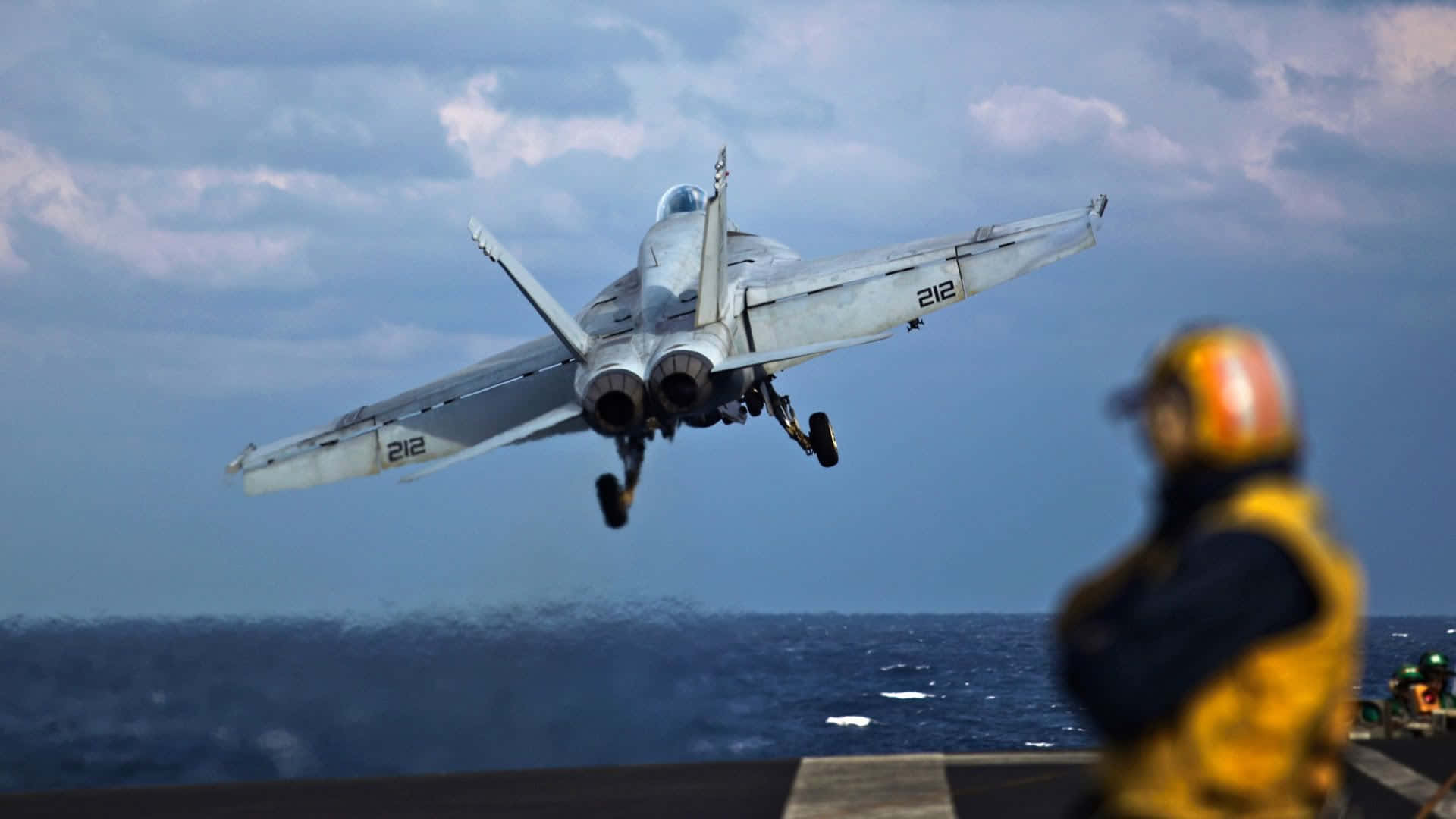 Navy 桌布 壁紙 — 一架灰色戰鬥機從航空母艦甲板起飛，下方有一名身穿黃色背心的船員在觀看。