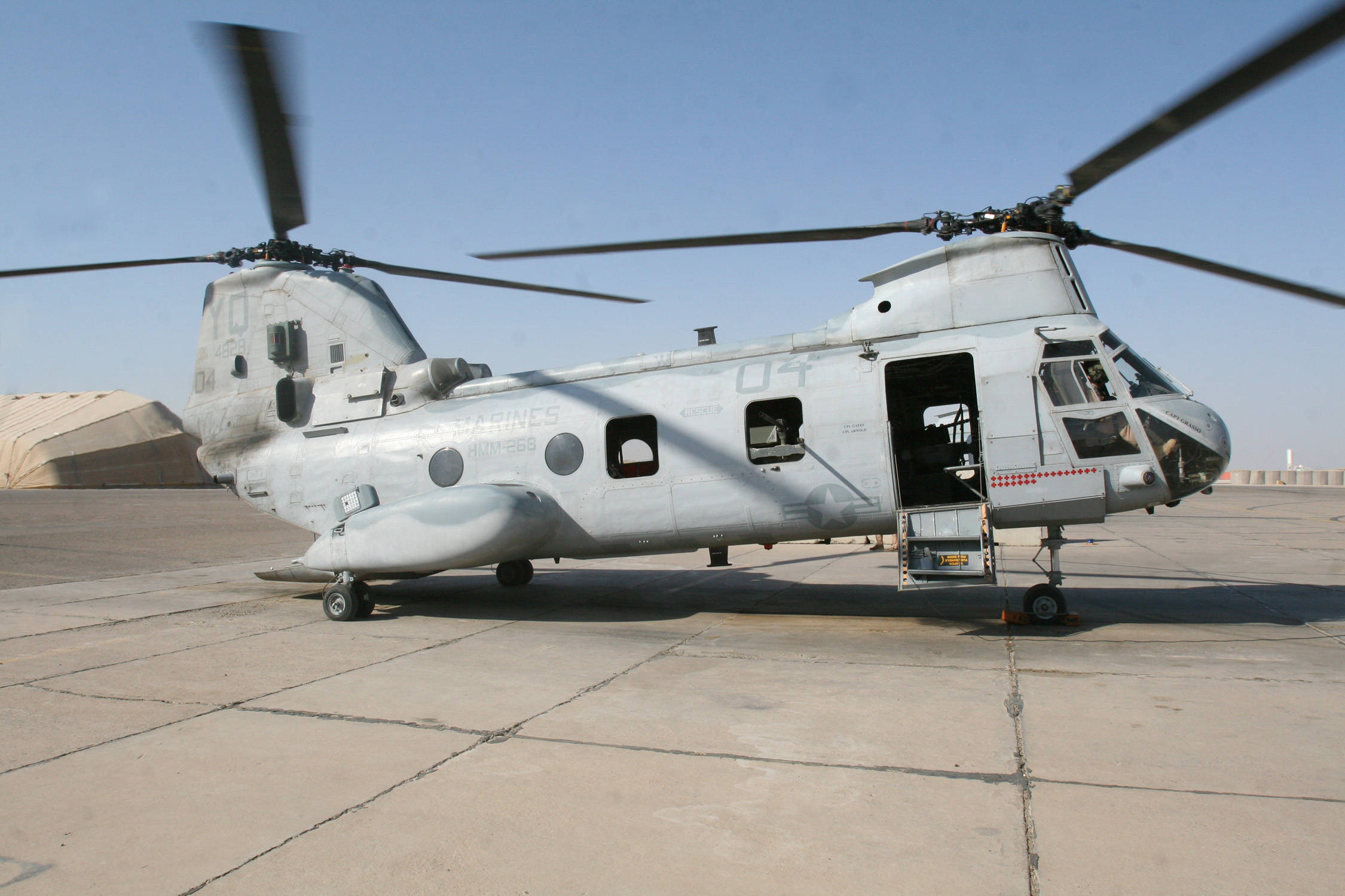 Sea Knight Military Helicopter 壁紙 — 一架灰色軍用直升機側門打開，靜止在混凝土停機坪上，天空清澈湛藍。