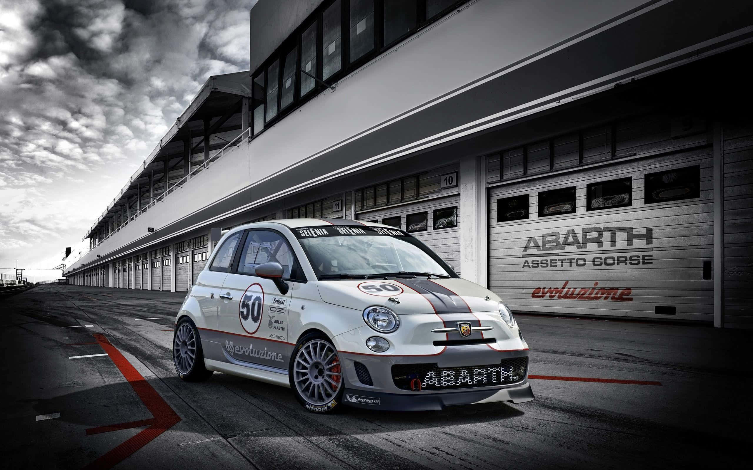 Abarth 桌布 壁紙 — 一架白色 Abarth 賽車，車號 50，停泊在賽車場車庫前，背景是戲劇性的多雲天空。
