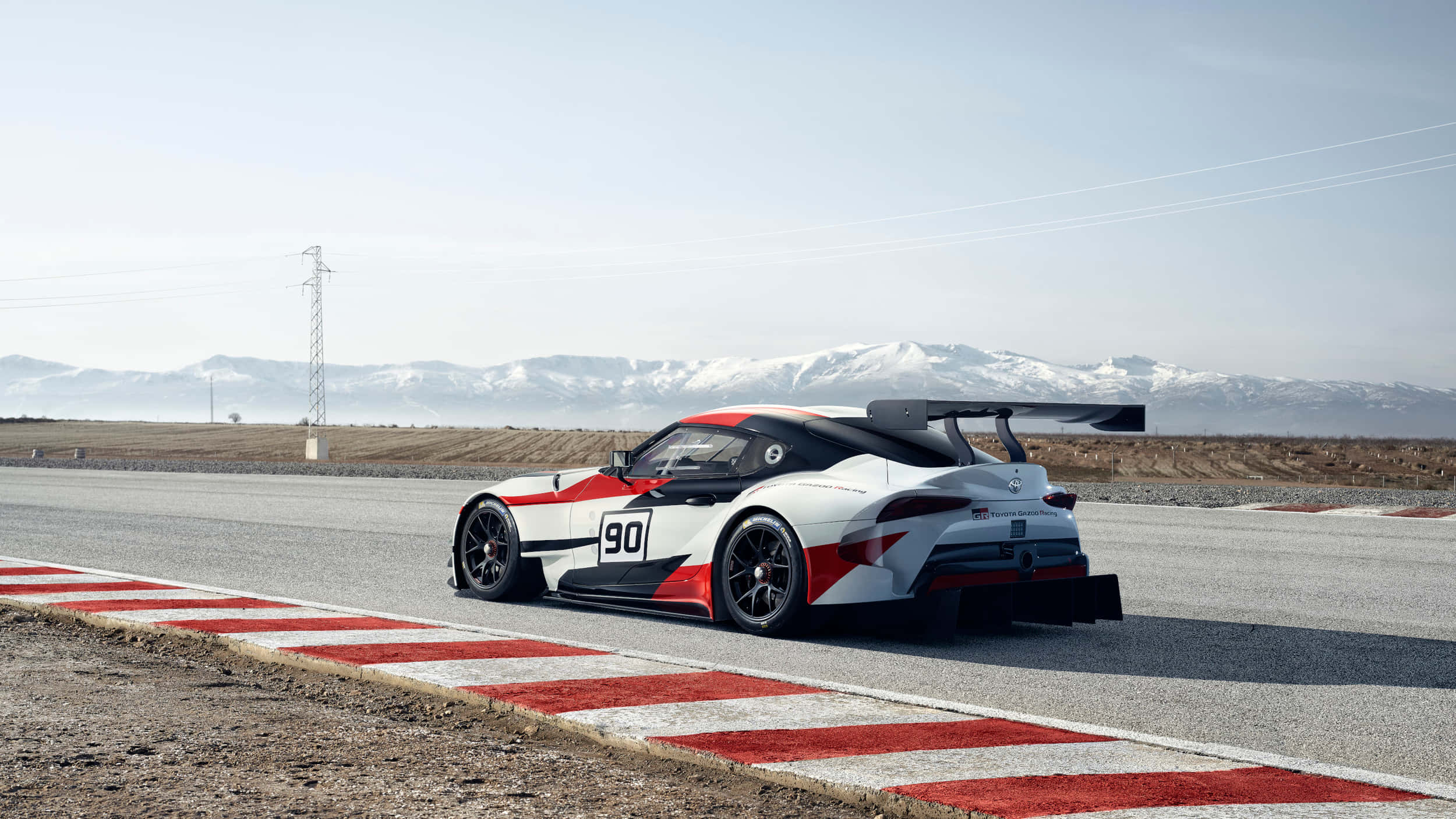 Supra 桌布 壁紙 — 一架白色、紅色和黑色、編號90的賽車，沿著由紅色和白色路緣石圍繞的賽道疾馳，遠處是白雪覆蓋的山脈。