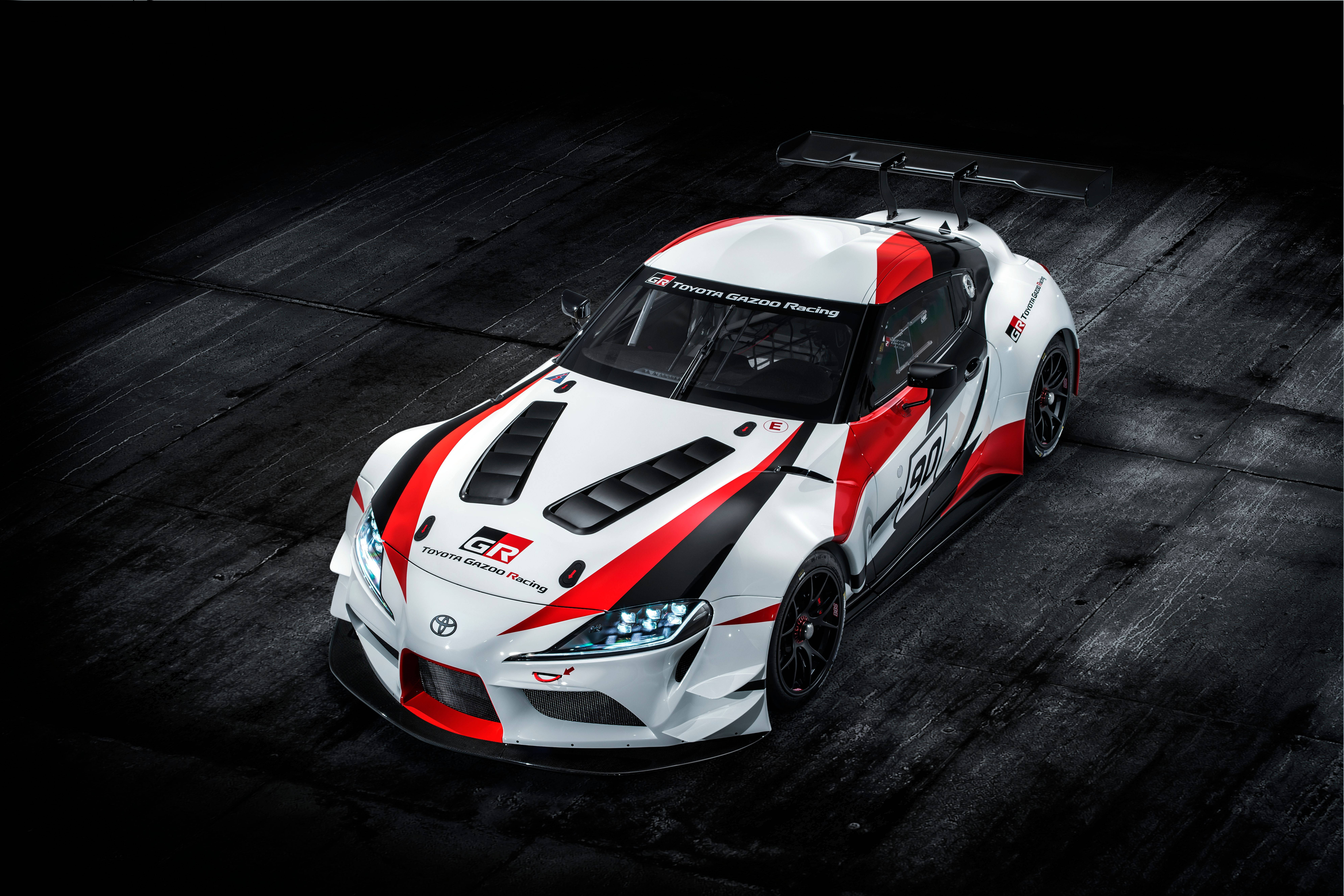 Gazoo Racing 4K Supra 壁紙 — 一架白色、紅色和黑色的豐田GR Supra賽車，帶有大型後擾流板，放置在深色木地板上，在戲劇性的工作室燈光下。